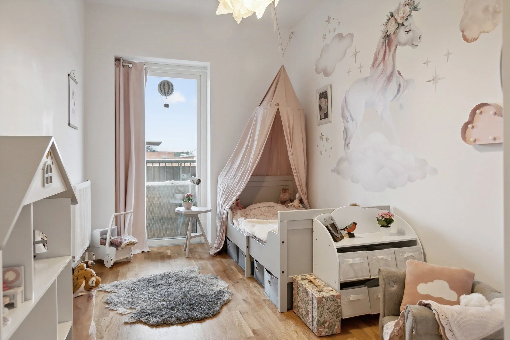 A cozy child's bedroom with a bed under a pink canopy, decorated with stuffed animals, and a large mural of a unicorn on the wall. The room features a sliding glass door leading to a balcony, soft furnishings, and pastel-colored decor.