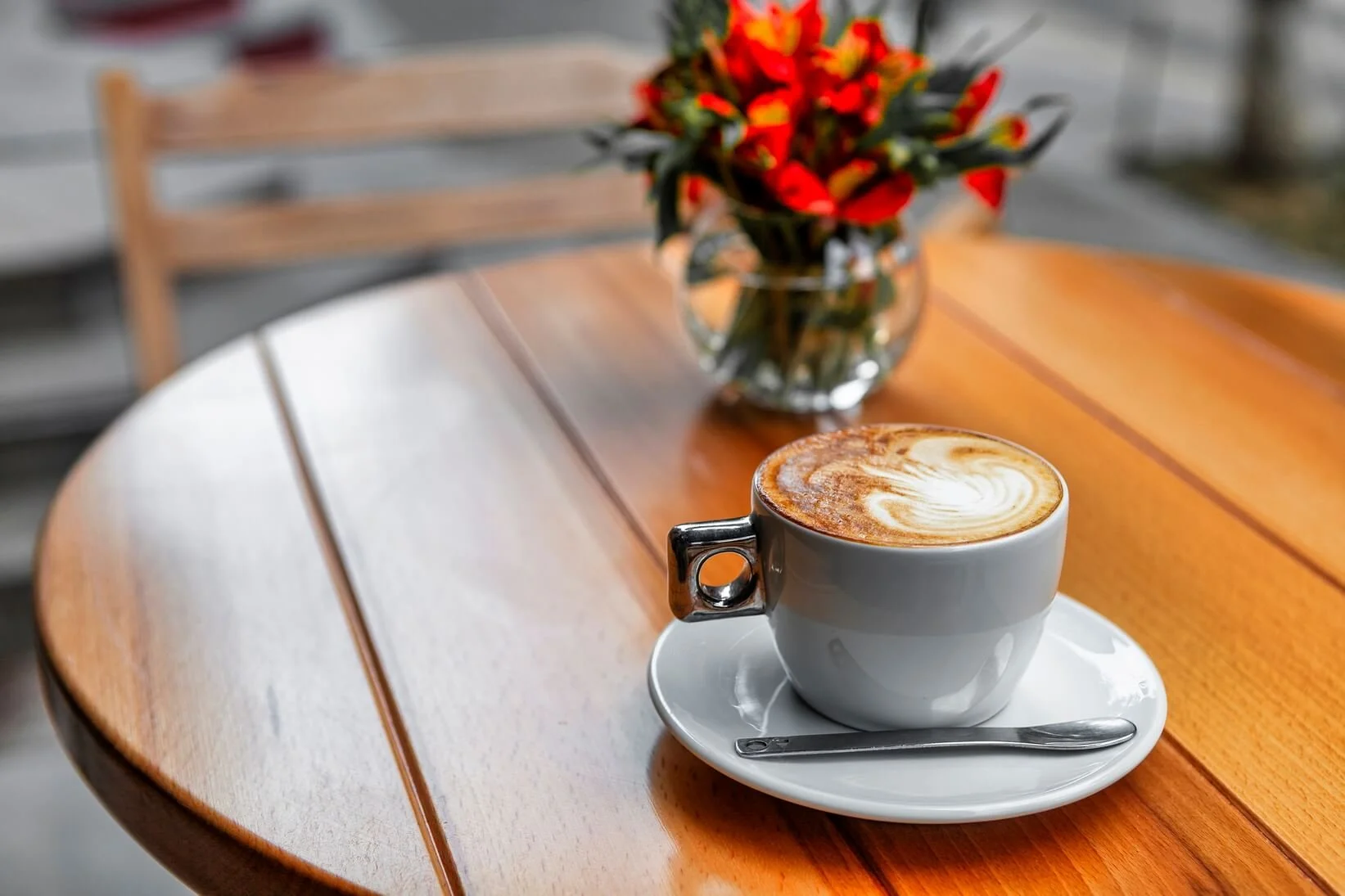A white ceramic cup of latte with foam art, placed on a saucer with a spoon on a wooden table, with a glass vase containing red and yellow flowers in the background.