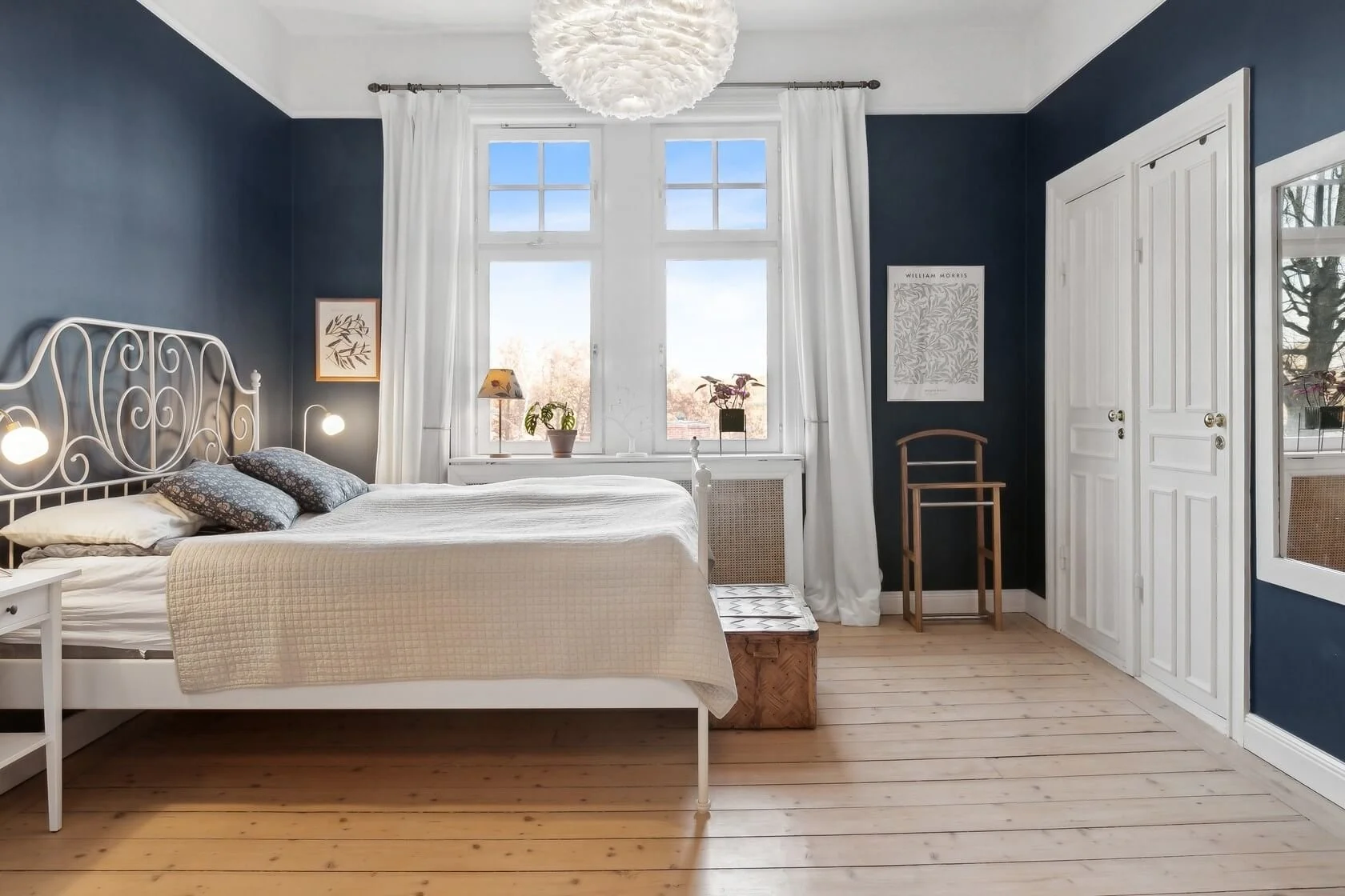 A cozy bedroom with dark blue walls, white bed frame and bedding, hardwood floors, large window with white curtains, nightstands with lamps, a framed picture on the wall, a standing mirror, a wooden chair, and a decorative chest at the foot of the be