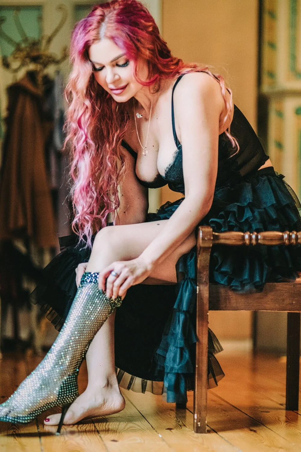A woman with long, curly pink hair sitting on a wooden chair while putting on a sparkling, high-heeled boot.