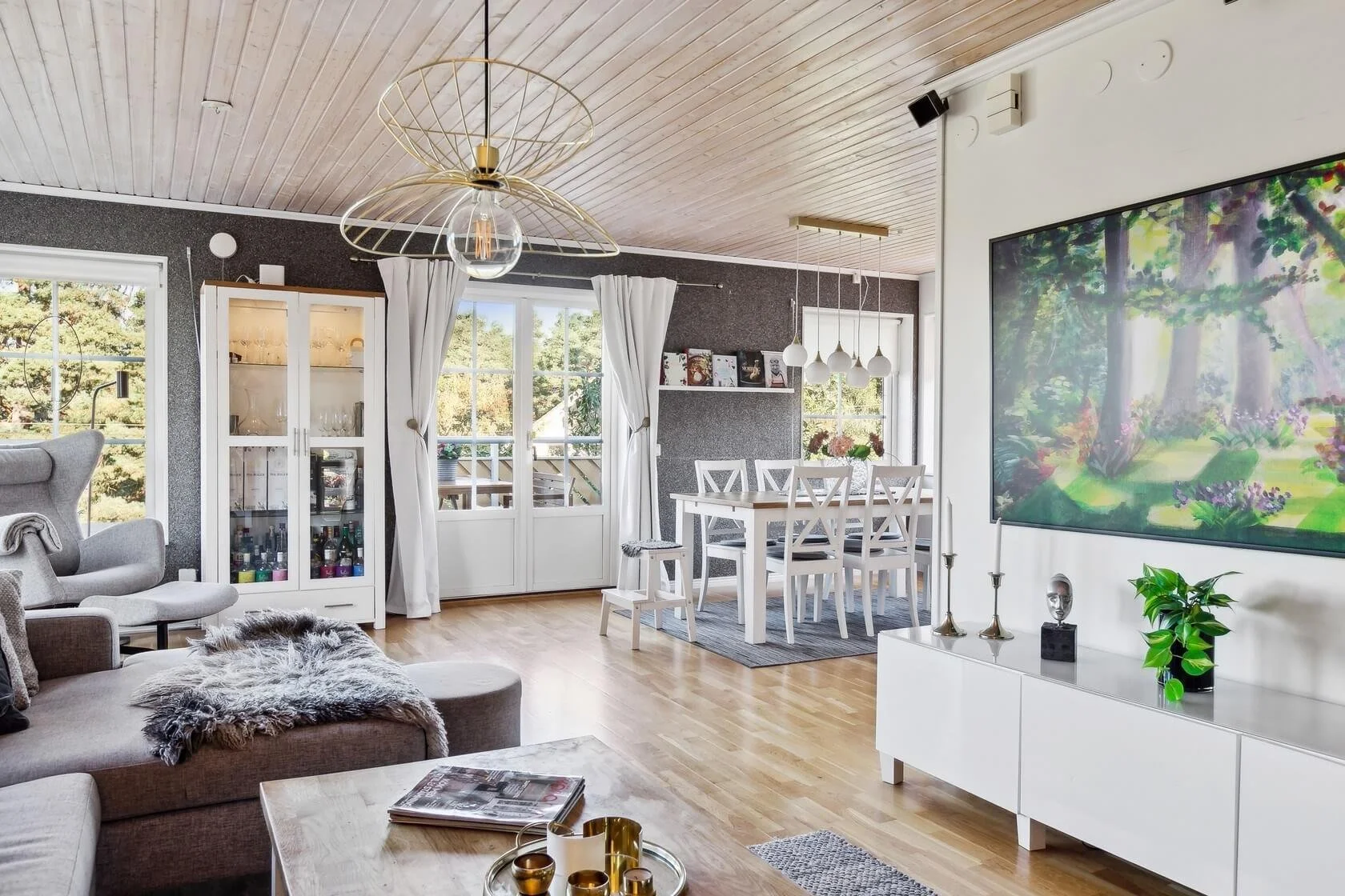 Bright living room with a dining area, large windows, white curtains, wooden flooring, modern furniture, and artwork on the wall.