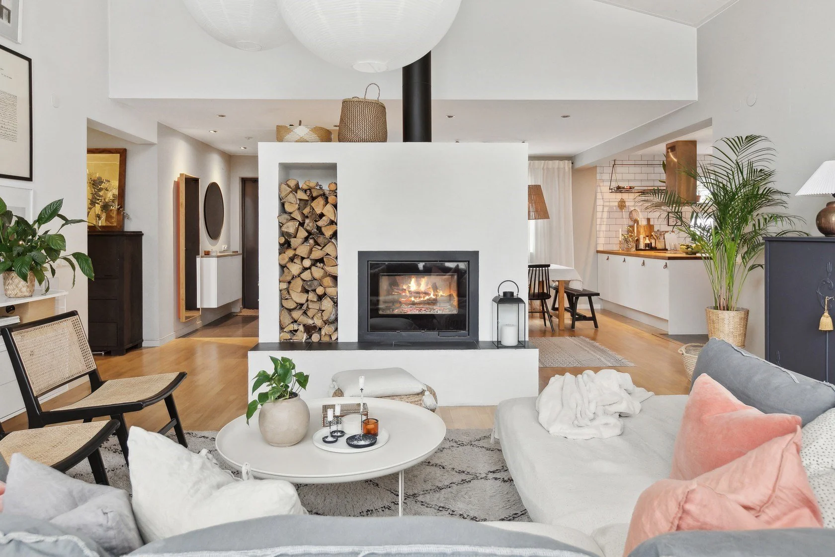 Living room with a white fireplace, wood logs, and a cozy fire, surrounded by plants, chairs, and a sofa with pillows.
