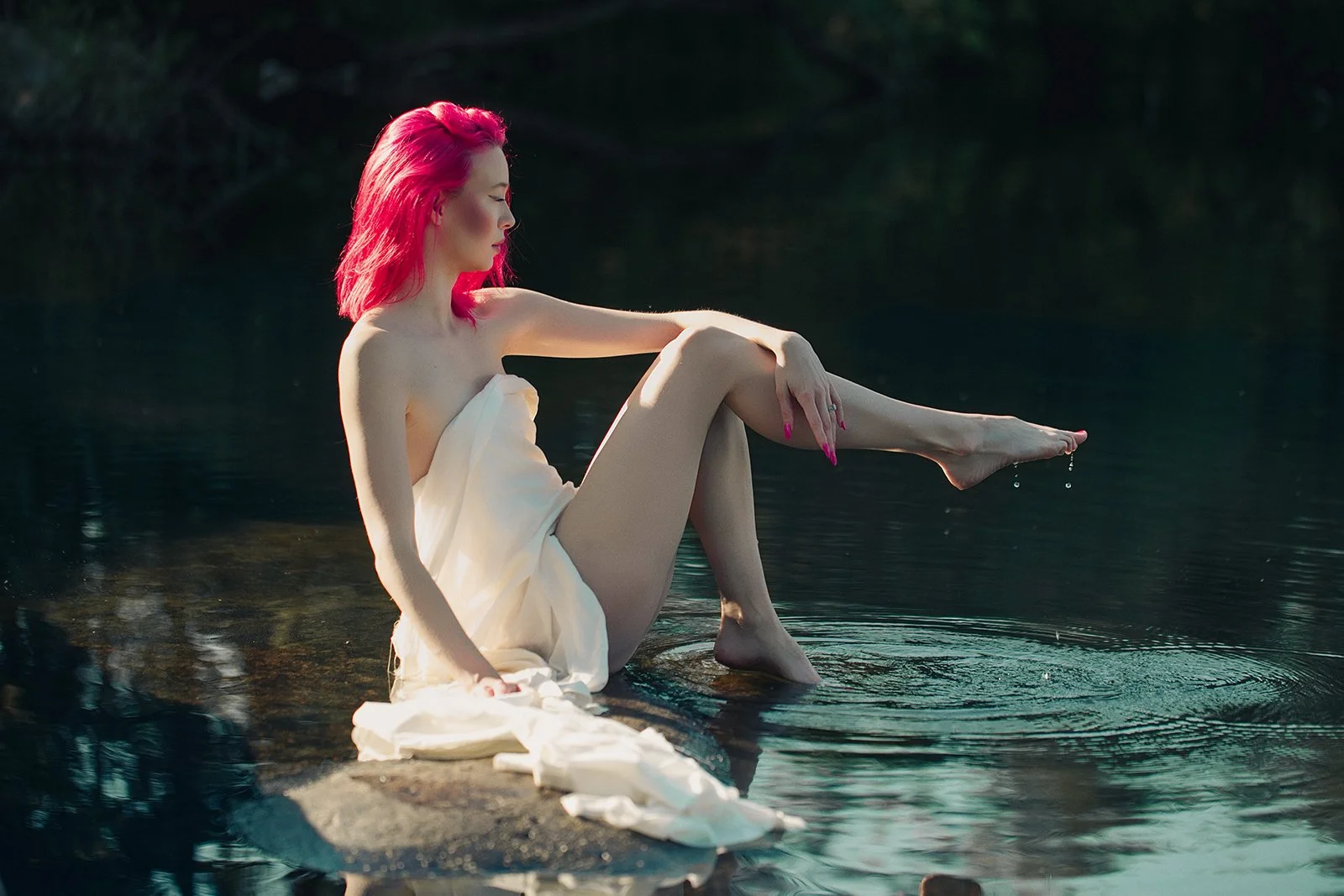 A woman with bright pink hair sitting on a rock by the water, wrapped in a towel, with her legs extended and draping fabric around her, as she dips her toes in the water.