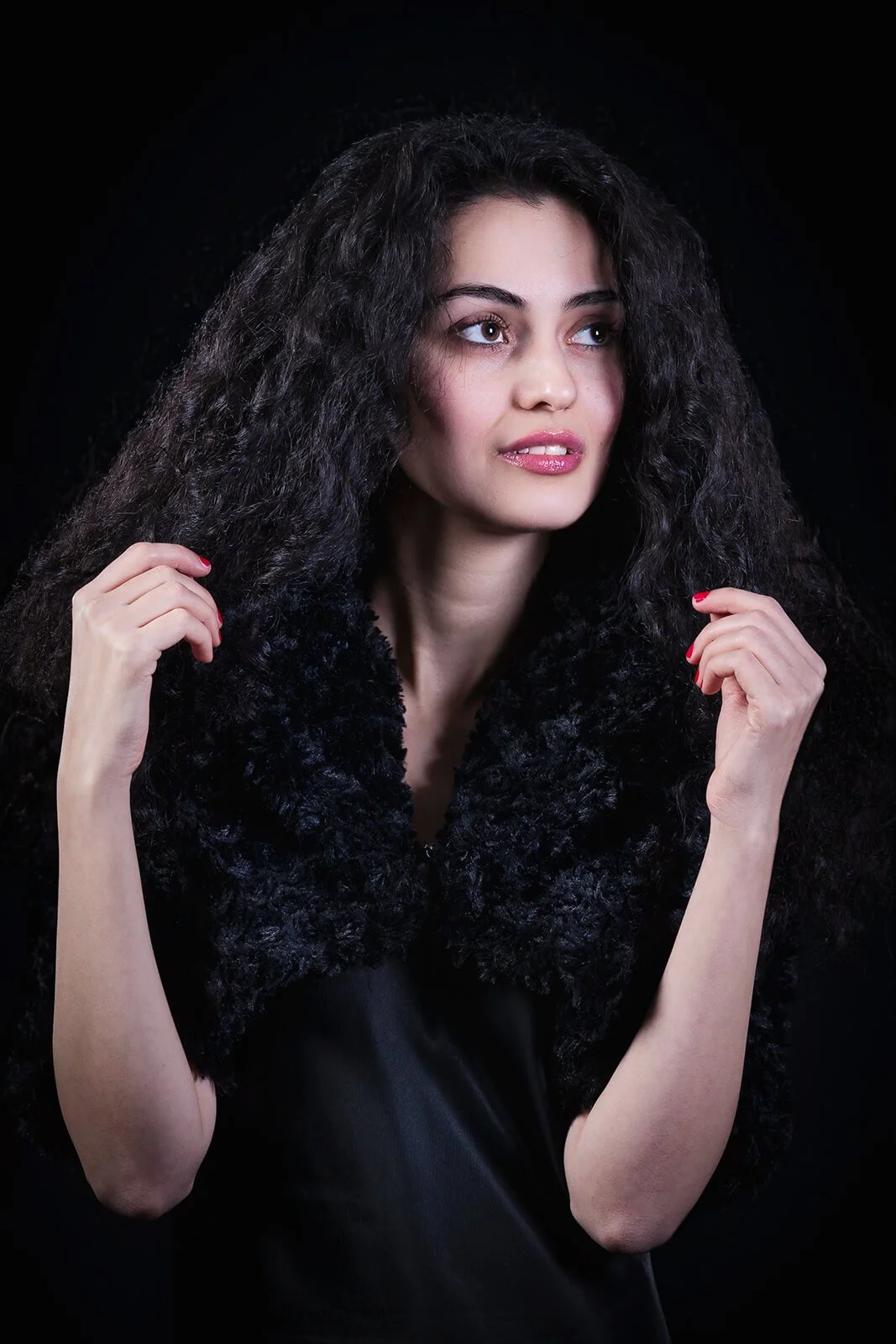 A woman with long, curly black hair, wearing a black fur shawl, looking to her right, against a dark background.