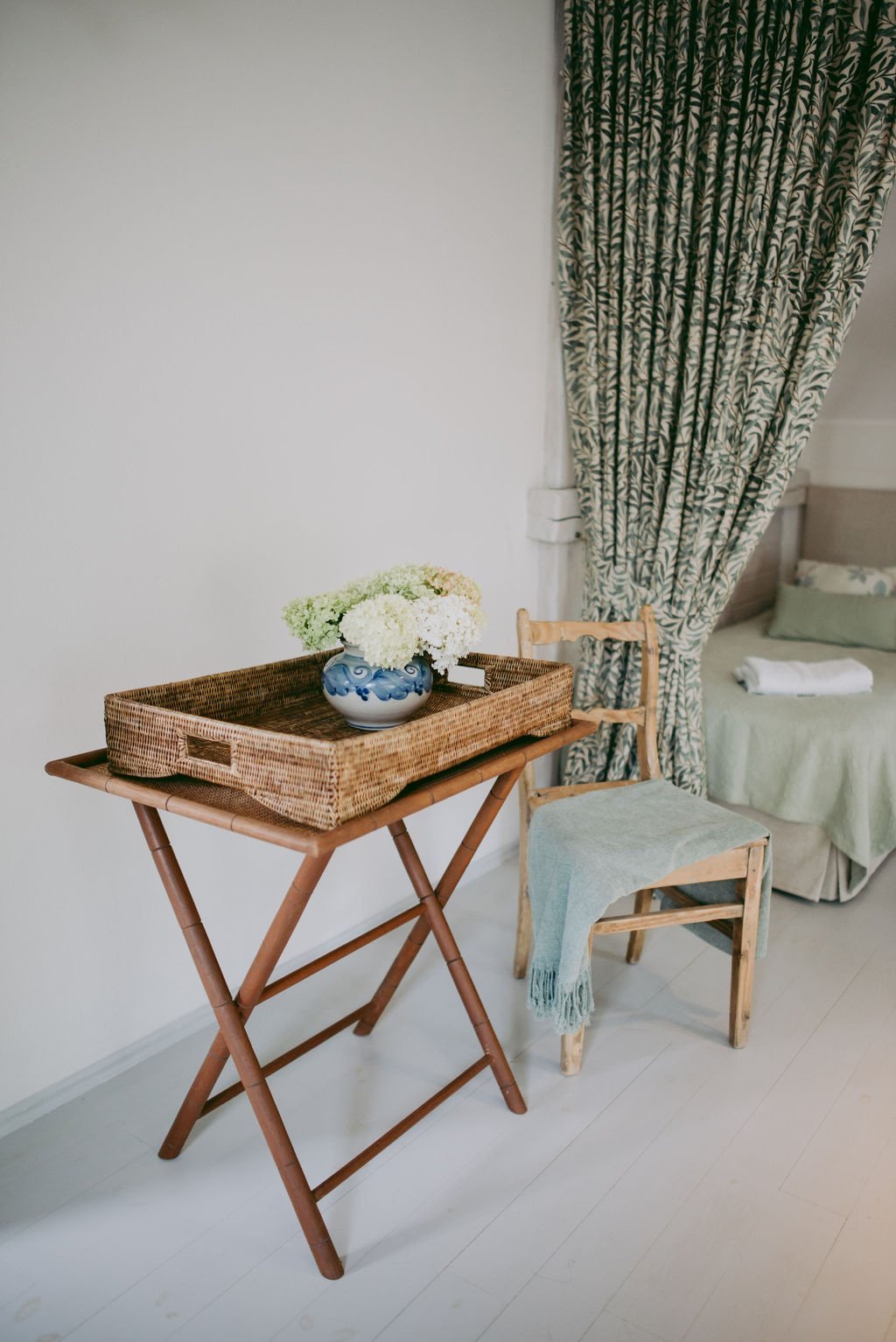 Interior scene with a wicker tray on a folding table, holding a blue and white vase with flowers, a wooden chair with a light green blanket, and a bed with green bedding and a towel.