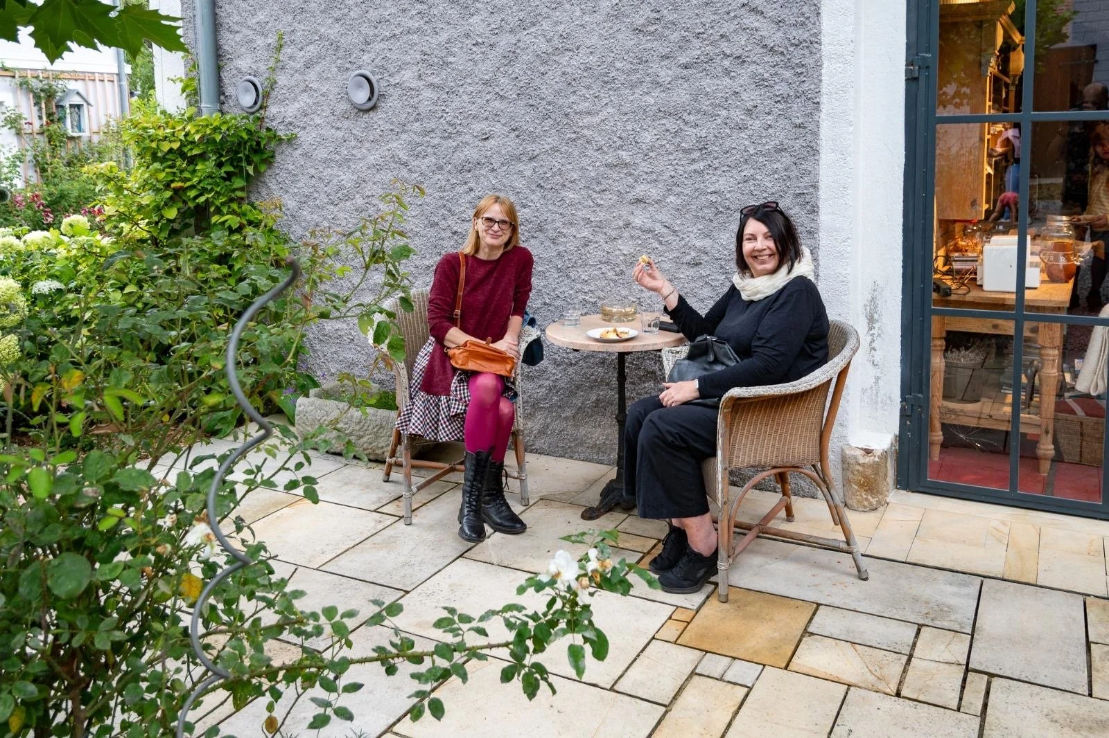 Two women sitting at an outdoor cafe table, smiling, surrounded by greenery and a stone wall.