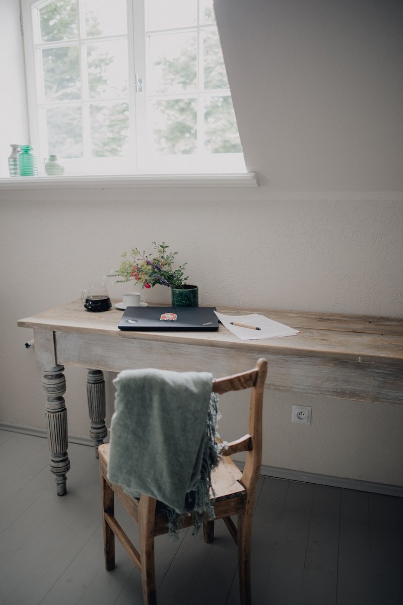 Rustic wooden desk with a laptop, papers, and a vase of flowers near a window. A chair with a draped blanket is in the foreground.