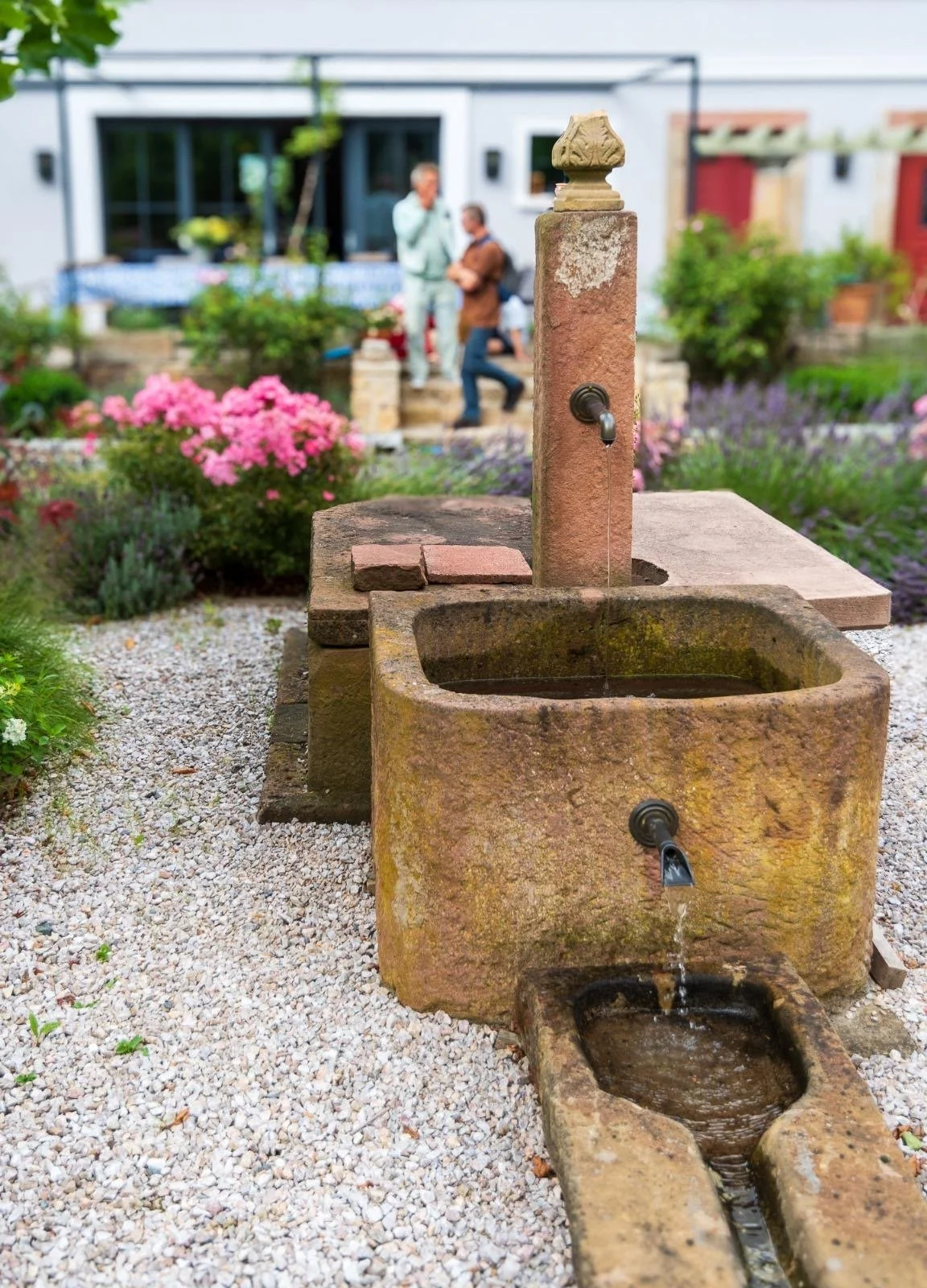Outdoor garden with stone fountain, flower beds, and two people in the background.