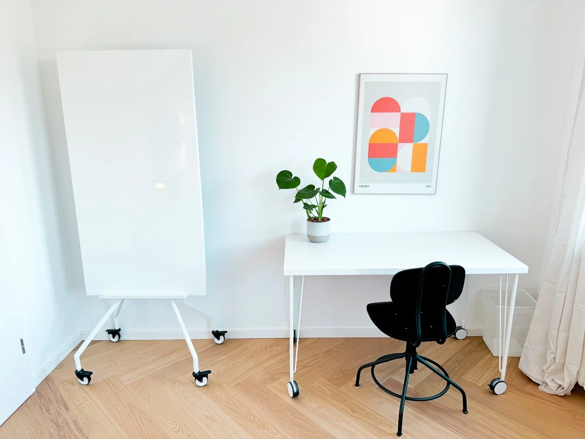 Ein modernes, minimalistisches Home-Office mit weißem Tisch, schwarzem Drehstuhl, Pflanzen und abstrakter Kunstdruck an der Wand