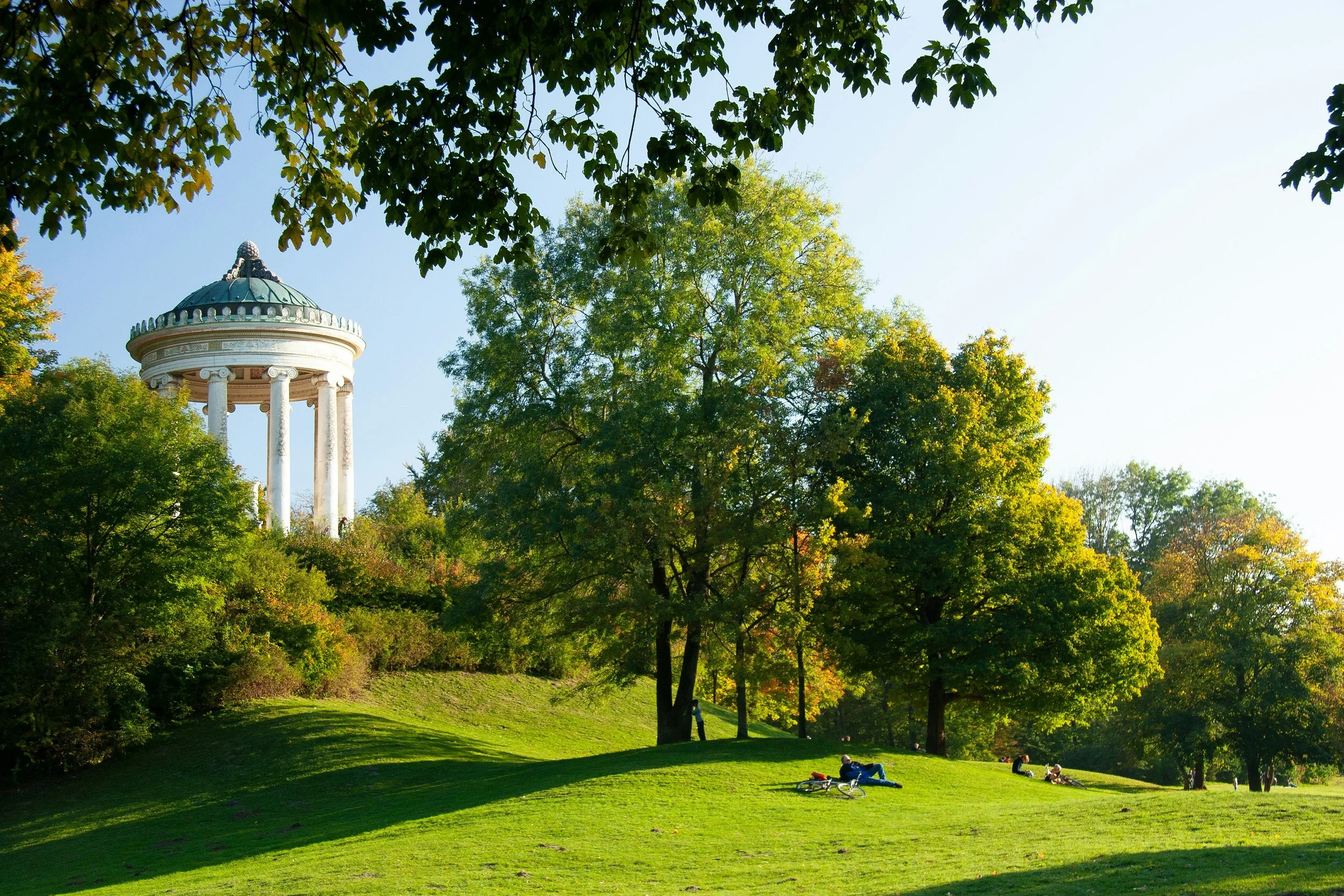 Grüner Park mit Menschen, die entspannen, und eine dorische Tempel-ähnliche Struktur auf einem Hügel im Hintergrund.