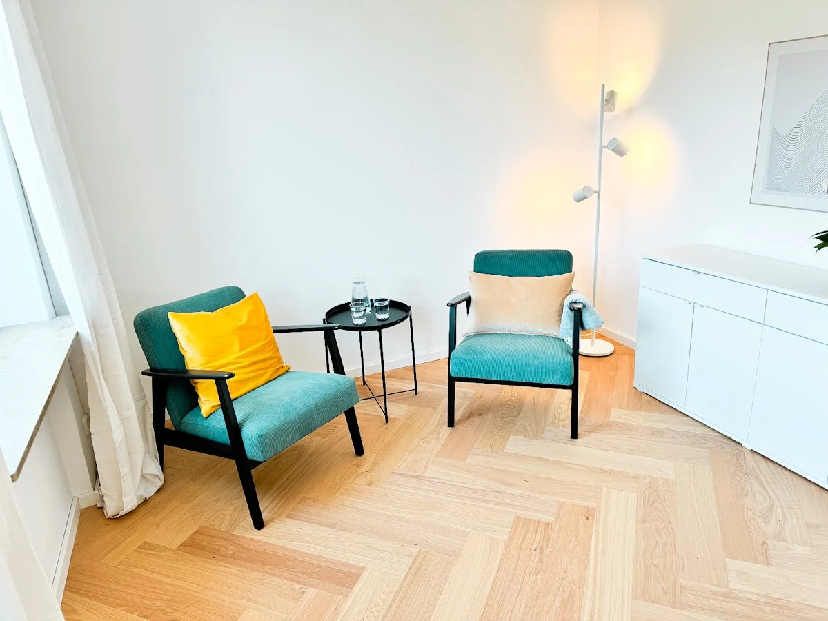 Zwei blaue Sessel mit Kissen, ein kleiner Beistelltisch mit Wasserflaschen, ein moderner Stehlampe, weiße Möbel und ein Bild an der Wand in einem hellen Wohnzimmer.