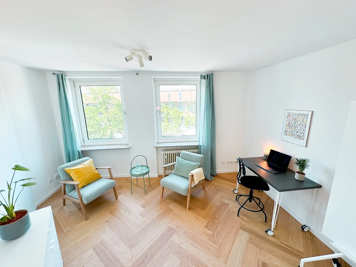 Gemütliches Wohnzimmer mit zwei grünen Sesseln, einem kleinen runden Tisch, einem Schreibtisch mit einem Laptop, einer Pflanze auf dem Tisch und einem Bild an der Wand.