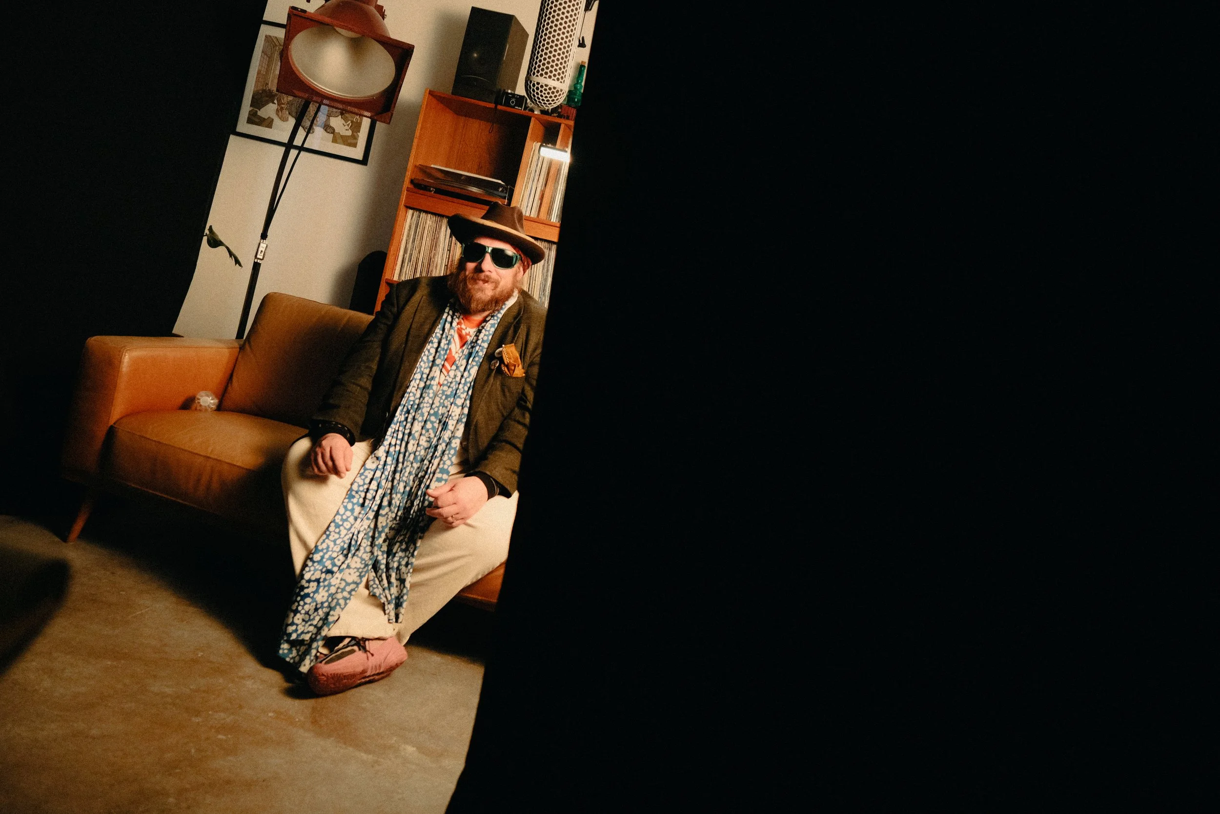 A man with a beard and sunglasses sitting on a tan sofa in a room with retro decor, including a tall lamp, records, and artwork on the wall.