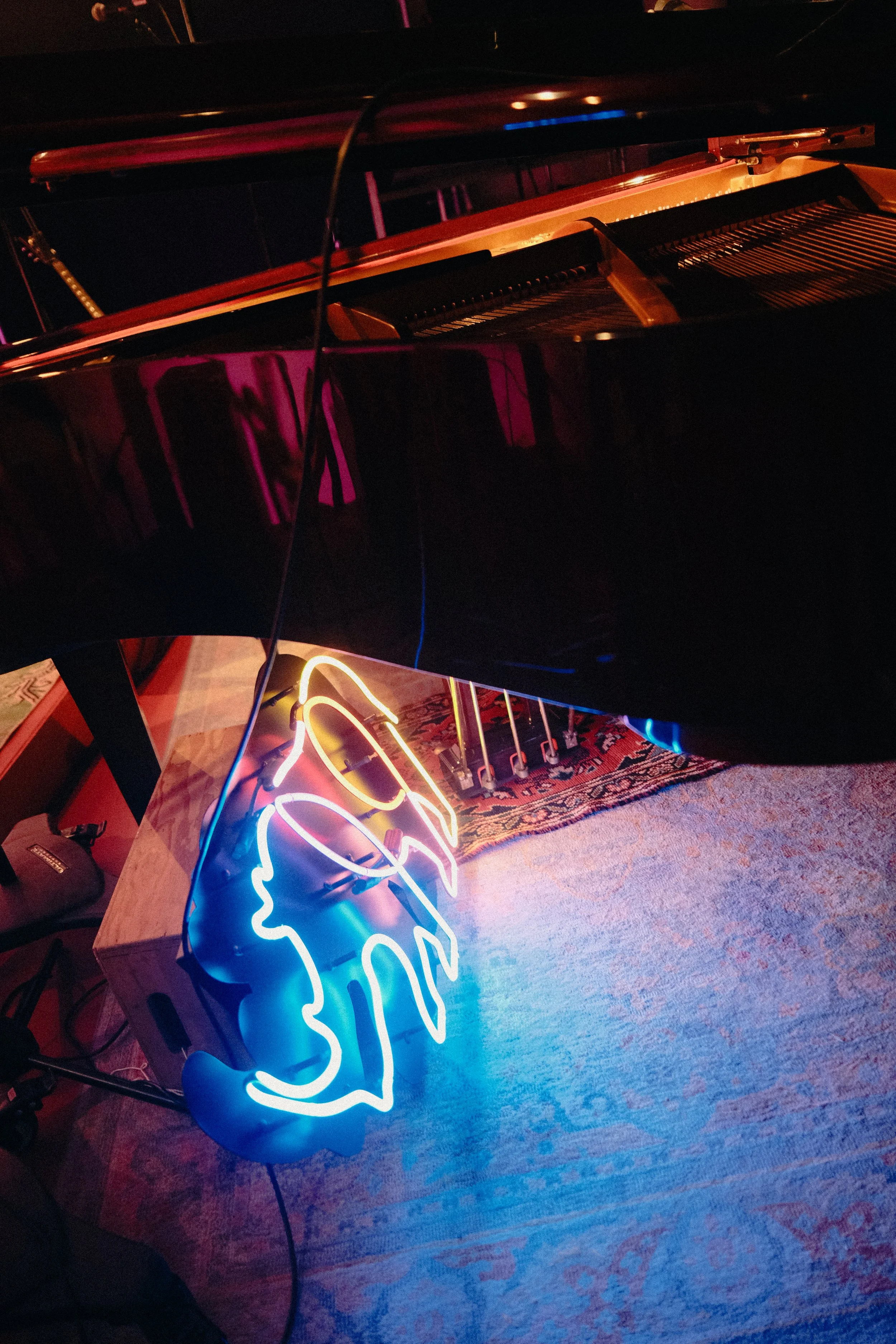 A grand piano with a neon sign of a dog's face with glasses hanging beside it, illuminated in blue and yellow, on a patterned rug in a dimly lit room.