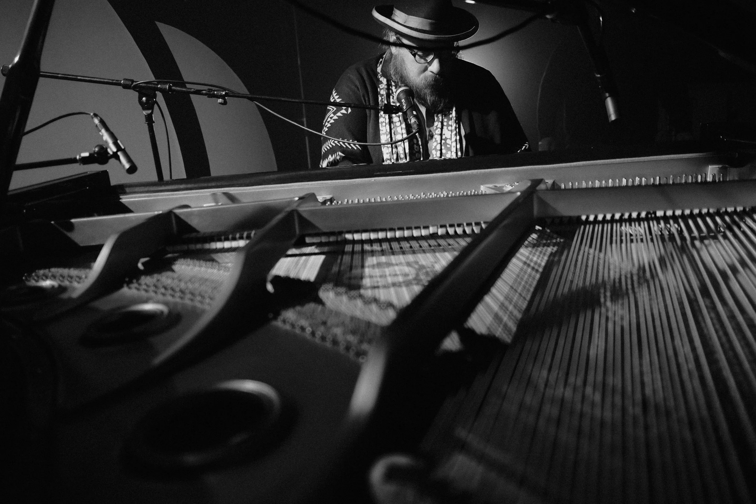 A musician wearing a hat, glasses, and patterned clothing playing a grand piano on stage with microphones above.