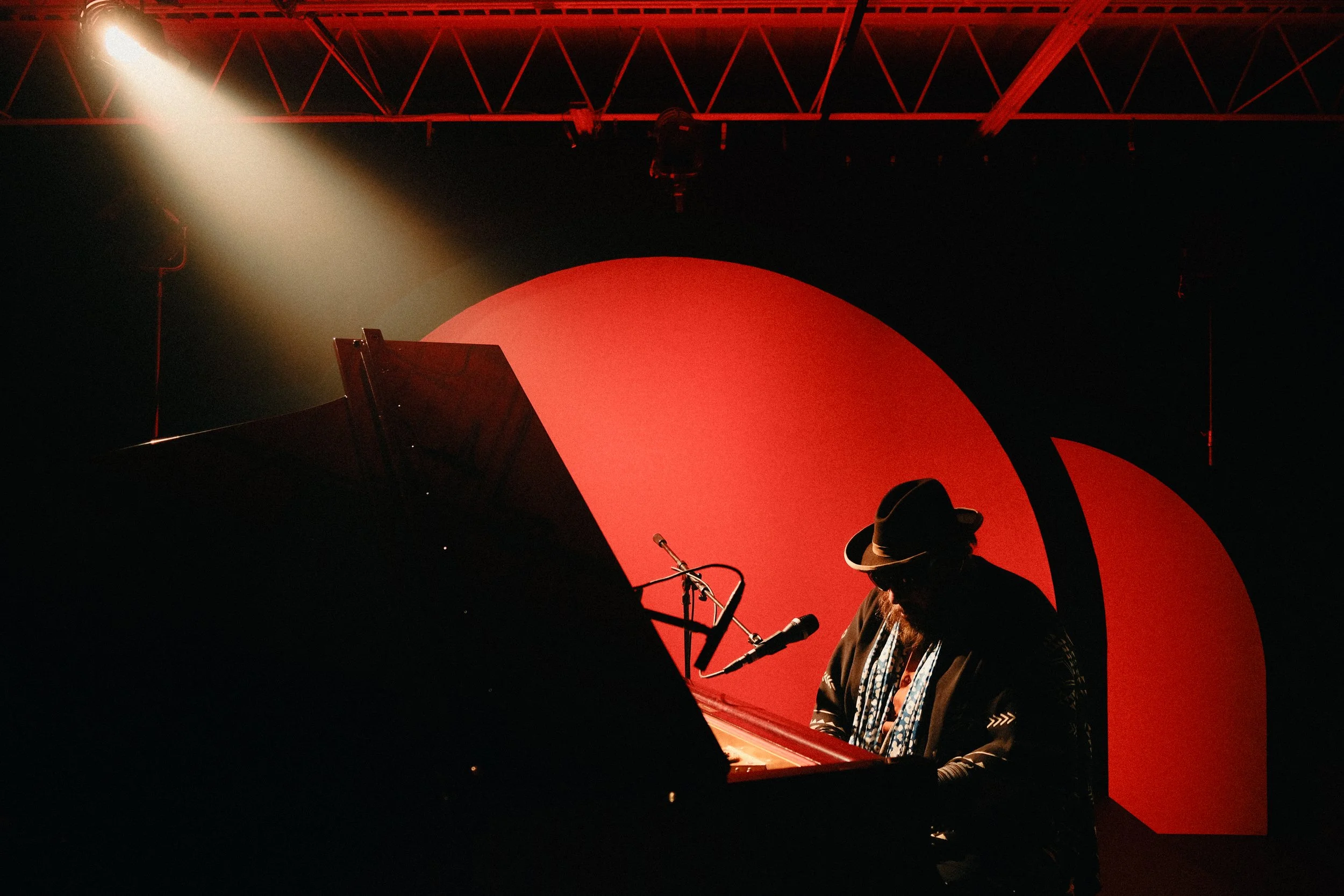 A person wearing a hat and glasses playing a grand piano in a dimly lit stage with a red circular backdrop and a spotlight.
