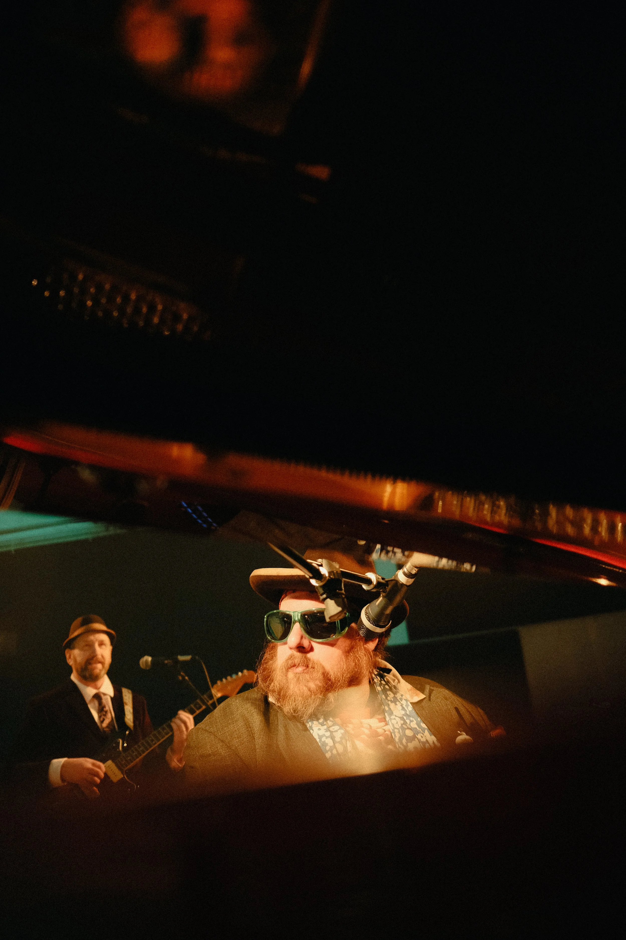 Two musicians performing on stage, one playing guitar and the other singing into a microphone, with the singer wearing sunglasses and a hat.