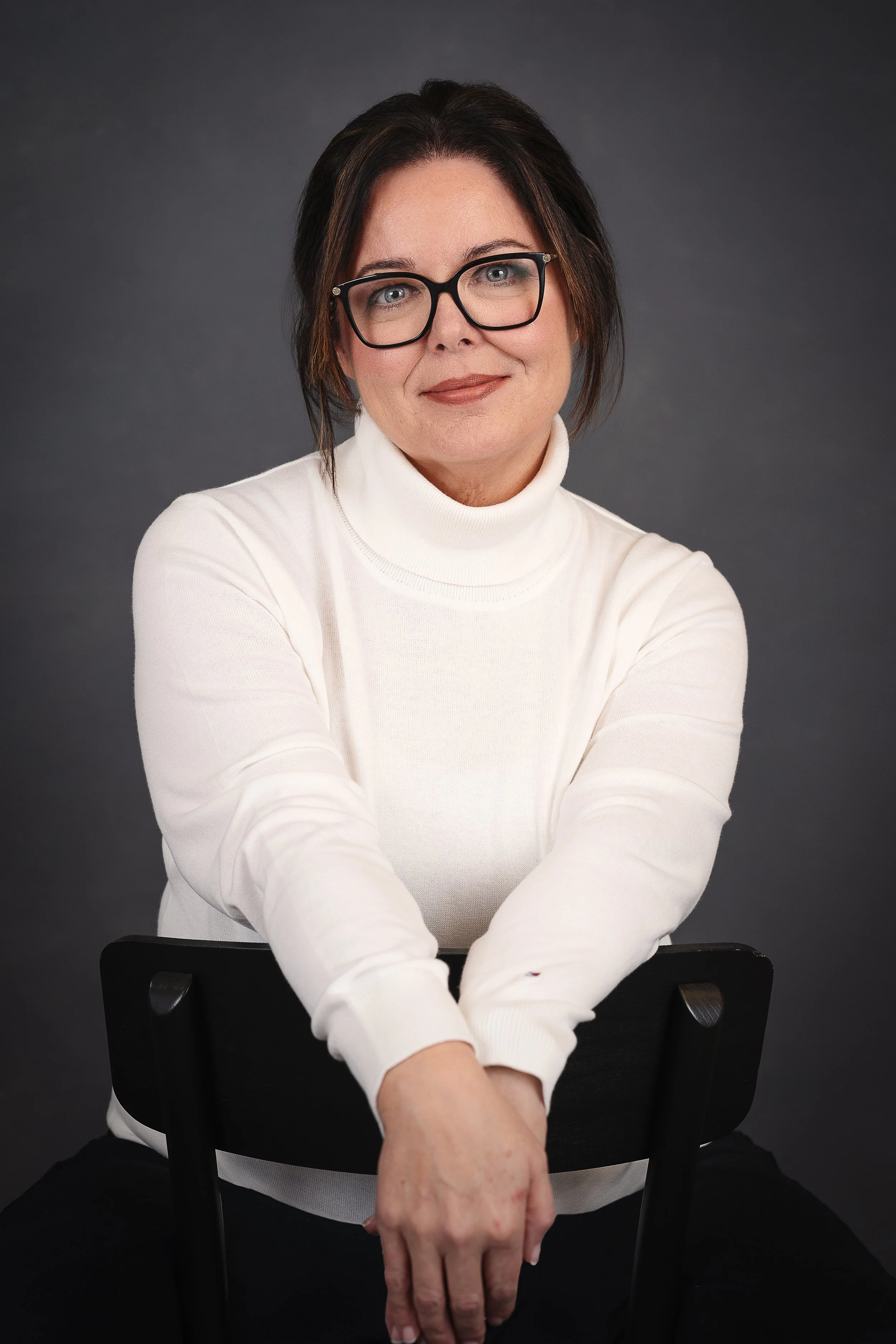 A woman with short dark brown hair, wearing glasses and a white turtleneck shirt, sitting on a black chair against a dark gray background. She is smiling slightly and has her arms resting on the back of the chair.