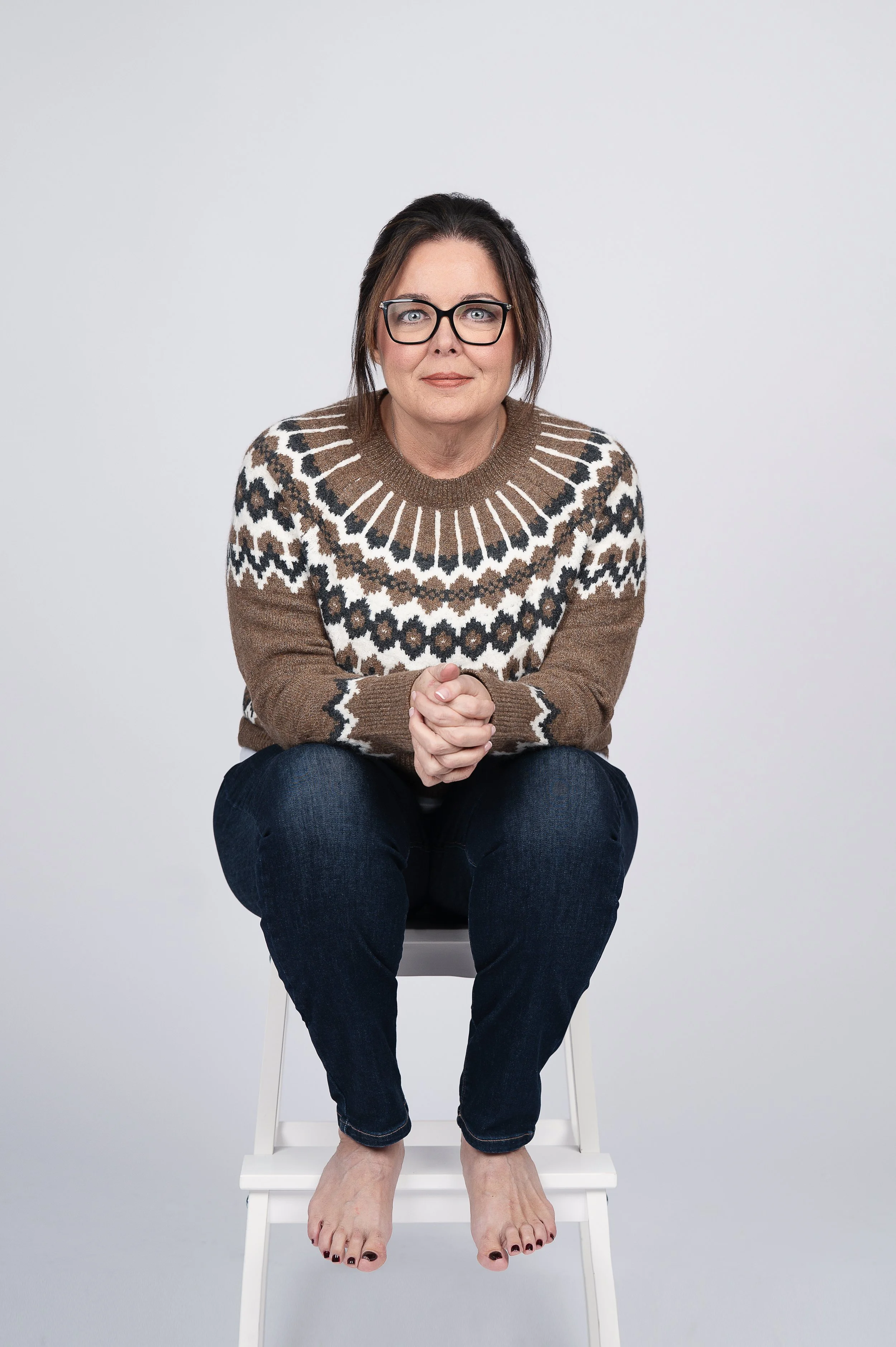 A woman with dark hair, glasses, and light skin sitting on a white chair against a plain gray background. She is wearing a brown, black, white, and beige patterned sweater and dark blue jeans, with her hands clasped together and her feet bare on the ground.