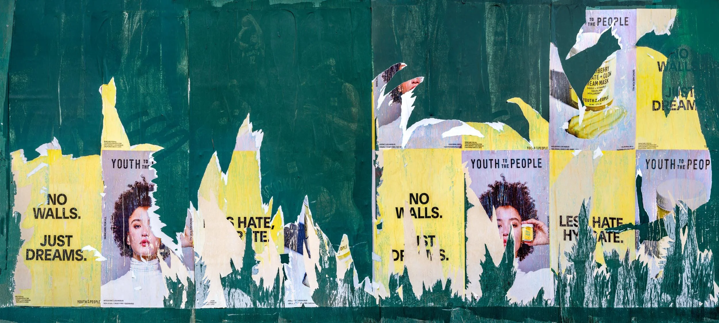 Fine art urban photography capturing decaying advertising posters on a plywood construction site fence. The composition isolates layers of shredded commercial paper, weathered textures, and accidental street art by Barton Lewis.
