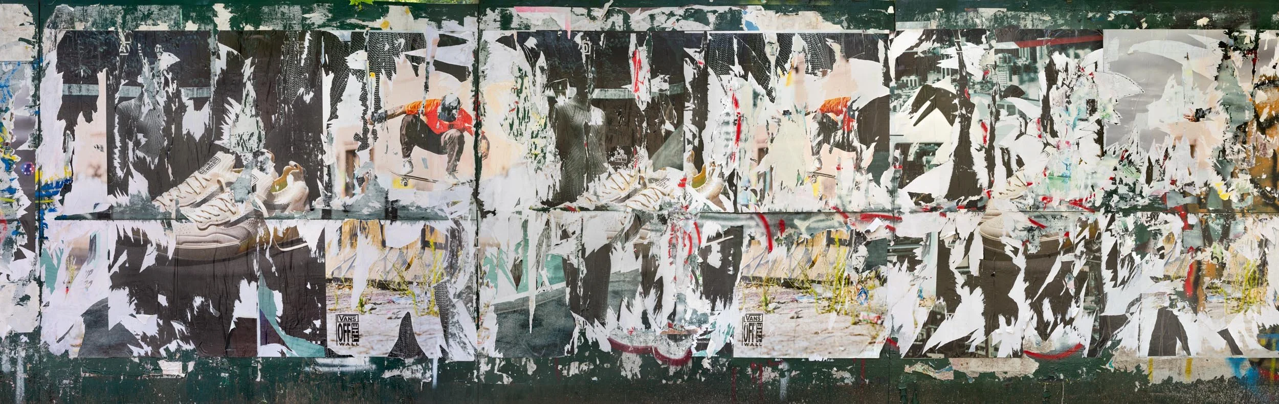 Fine art urban photography capturing decaying advertising posters on a plywood construction site fence. The composition isolates layers of shredded commercial paper, weathered textures, and accidental street art by Barton Lewis.