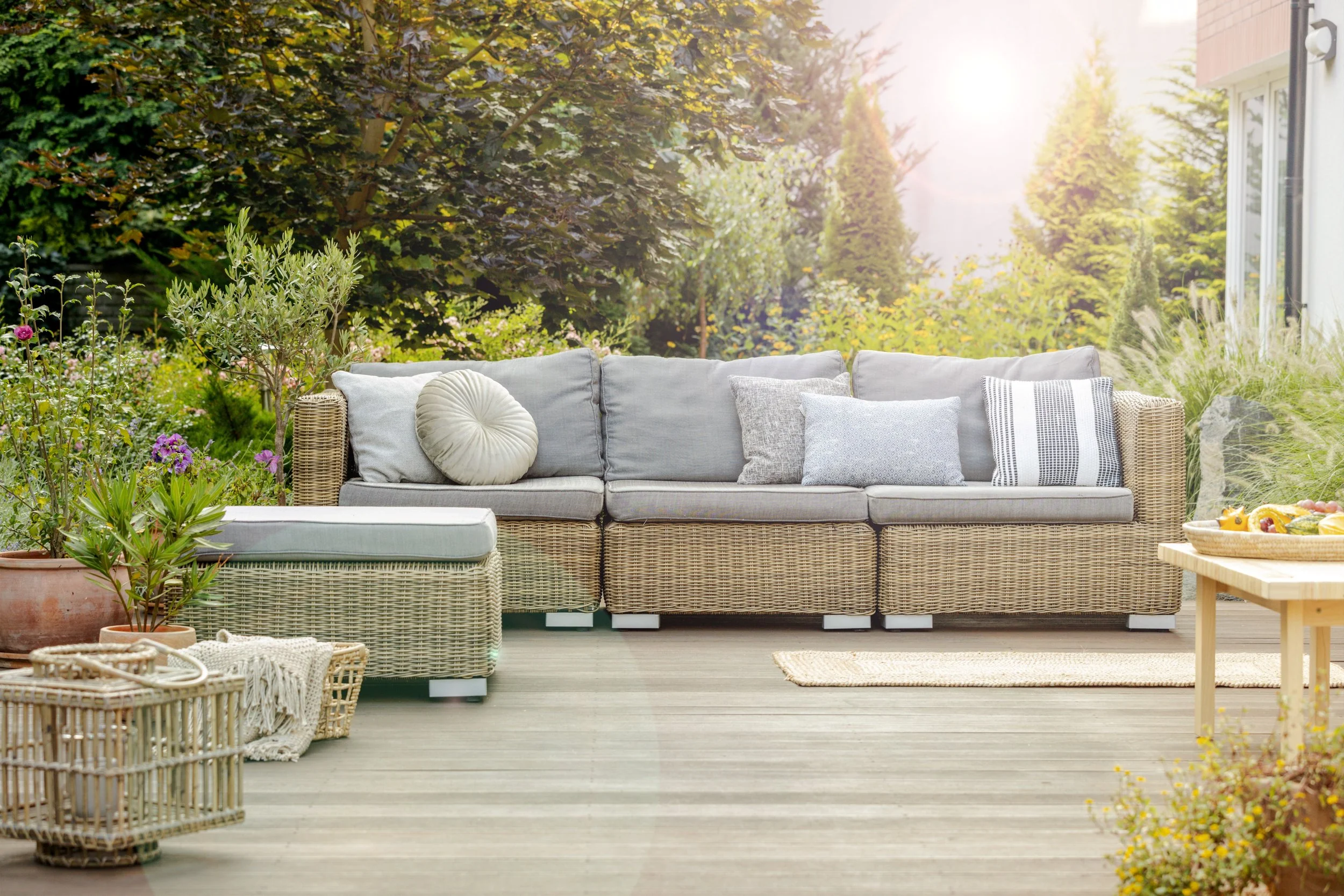 Outdoor patio with wicker sofa, various pillows, potted plants, a wicker ottoman, a basket with a blanket, and a table with a fruit bowl, surrounded by lush green trees and sunlight.