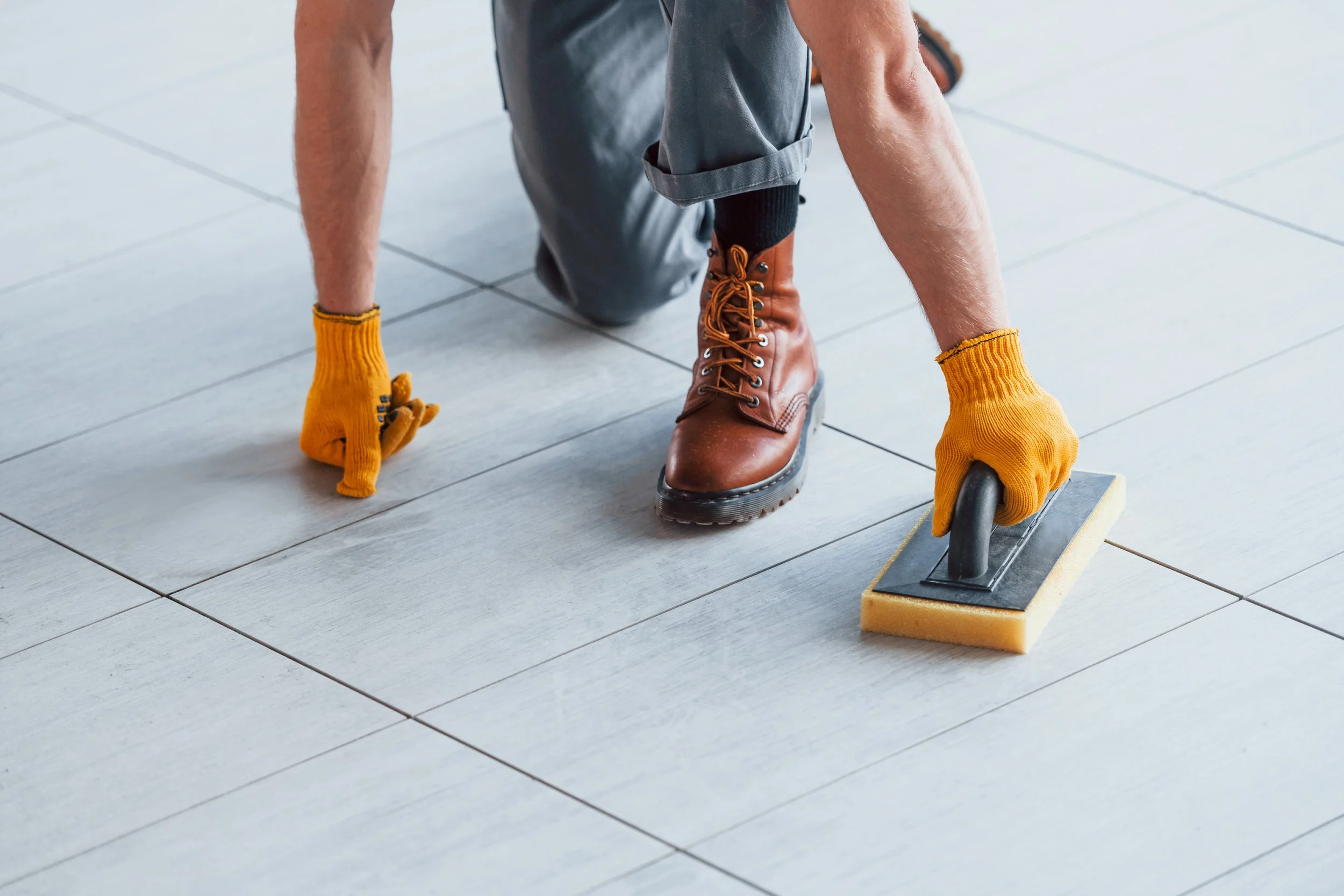 Person installing or repairing flooring using a hand trowel and sponge, wearing yellow gloves and brown boots.