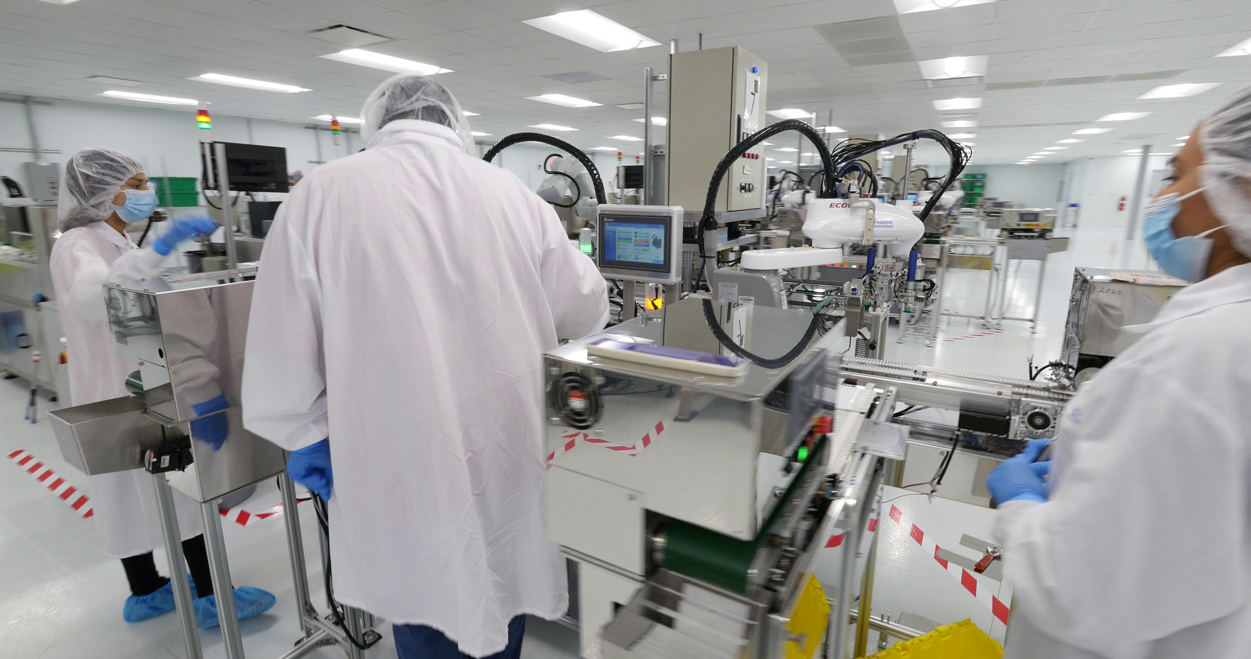 Scientists wearing lab coats, masks, and gloves working in a clean manufacturing facility with automated machinery.