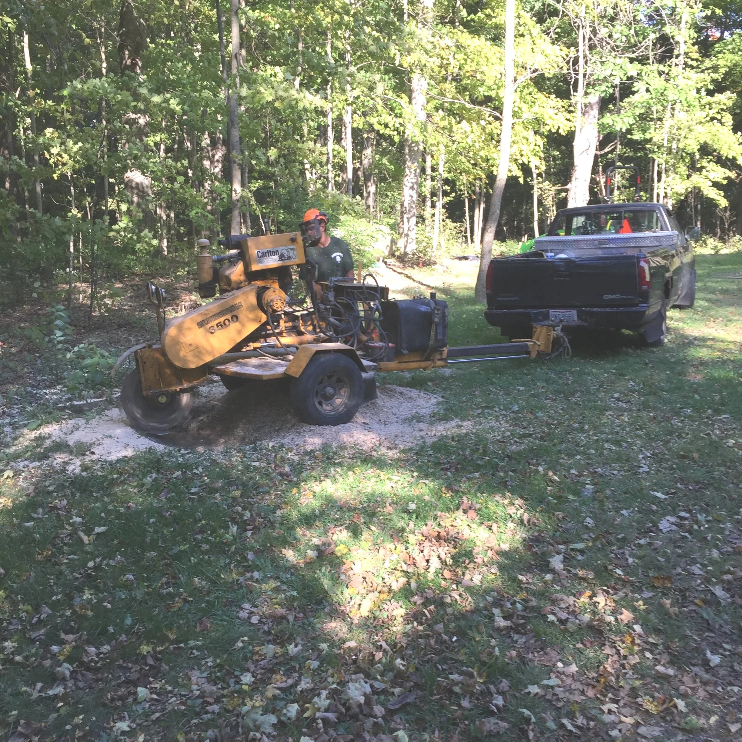 A person operating a yellow road milling machine on grass and gravel, connected to a black GMC pickup truck, in a wooded area with green trees.