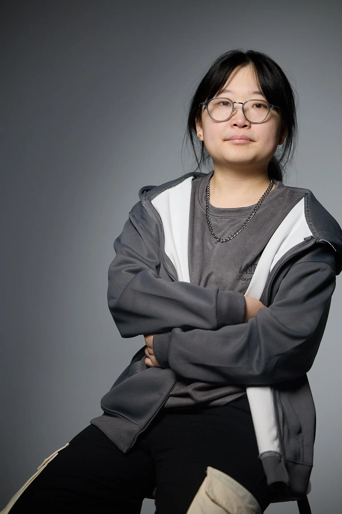 A woman with glasses and long brown hair, wearing a purple blazer over a black top, jeans, and sneakers, sitting on a stool with a neutral background.