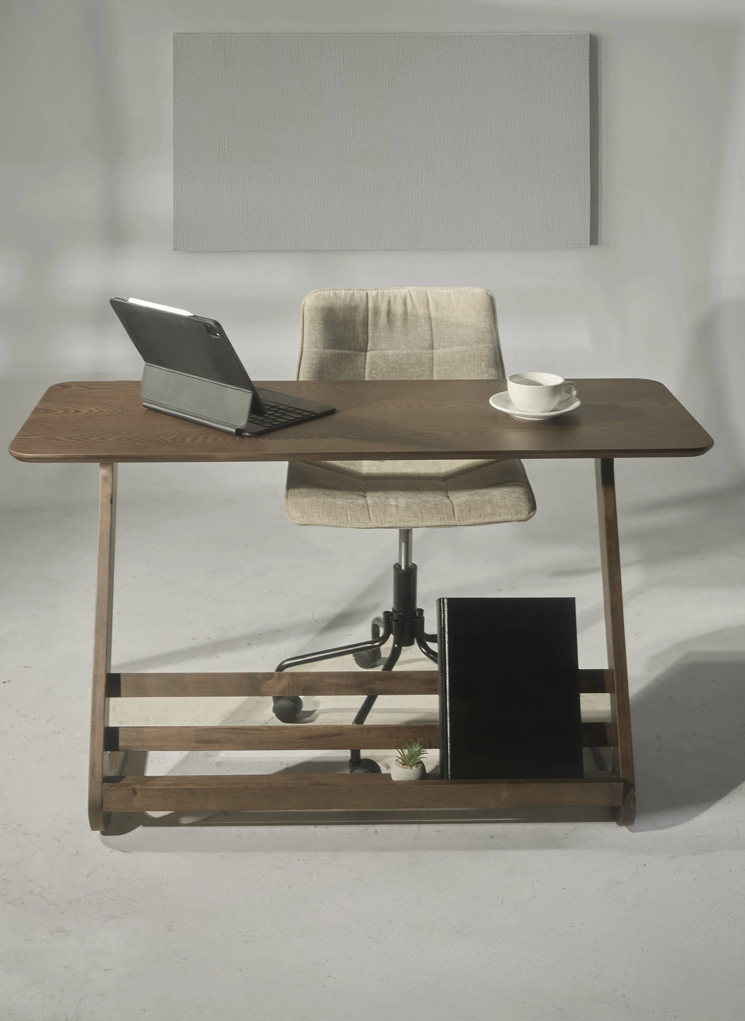 A wooden desk with an open tablet on a stand, a white coffee cup on a saucer, a black binder, and a small potted succulent plant. An office chair behind the desk and a large white wall with a gray acoustic panel in the background.
