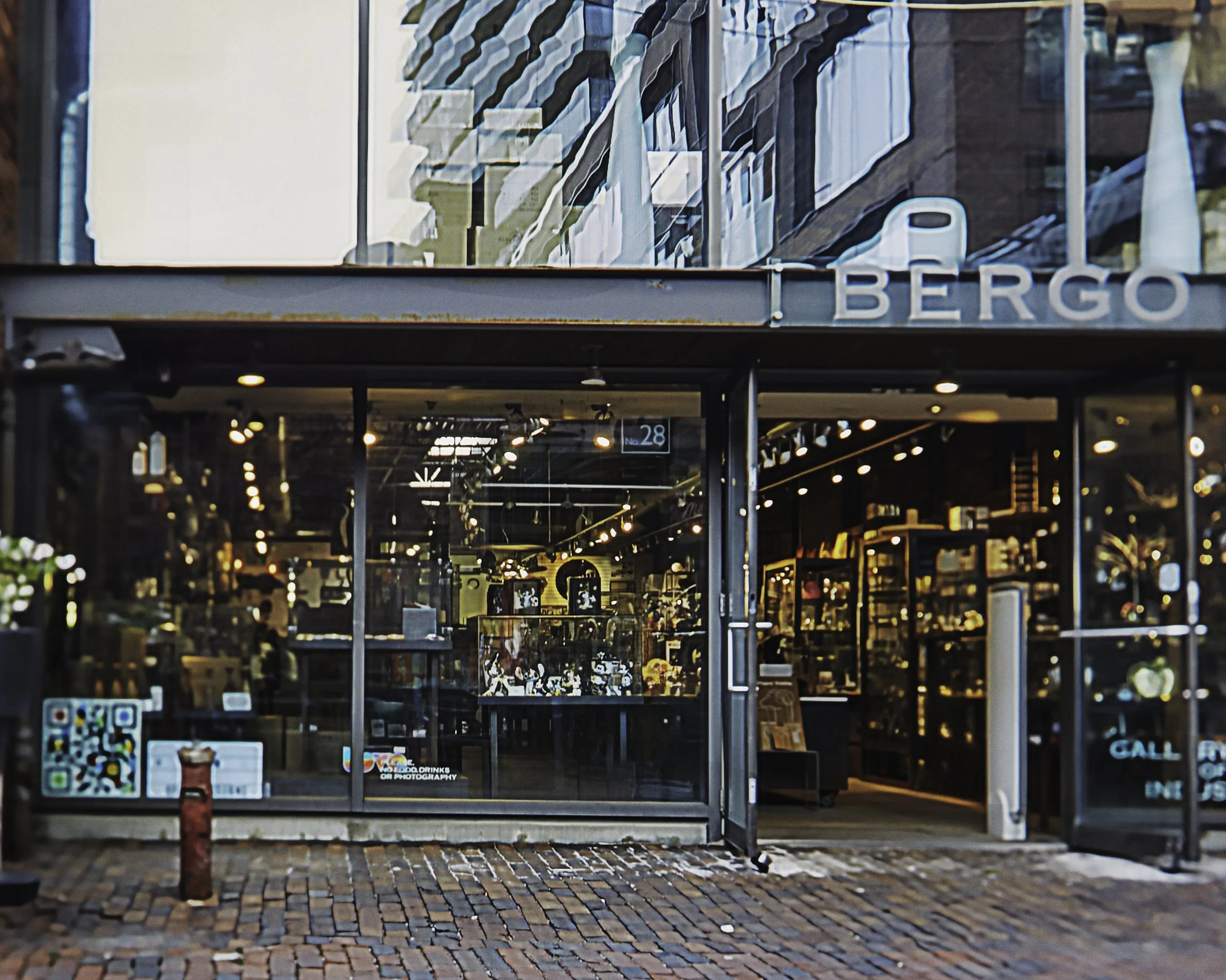 Front view of a modern retail store named 'Bergo' with large glass windows, displaying lights and shelves filled with products inside. The entrance is open, showing part of the interior with warm lighting and merchandise.