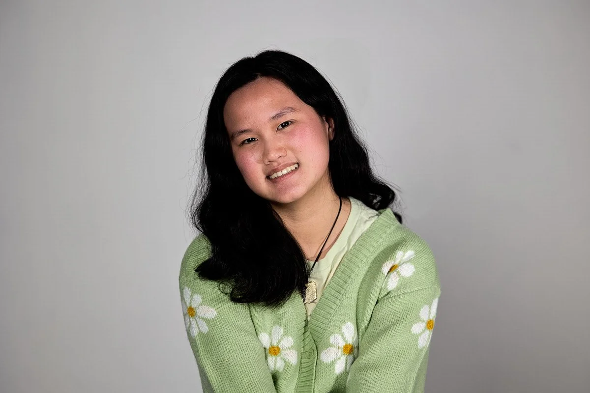 Young woman with long black hair wearing a light green sweater with white and yellow daisies, smiling and looking at the camera against a plain gray background.