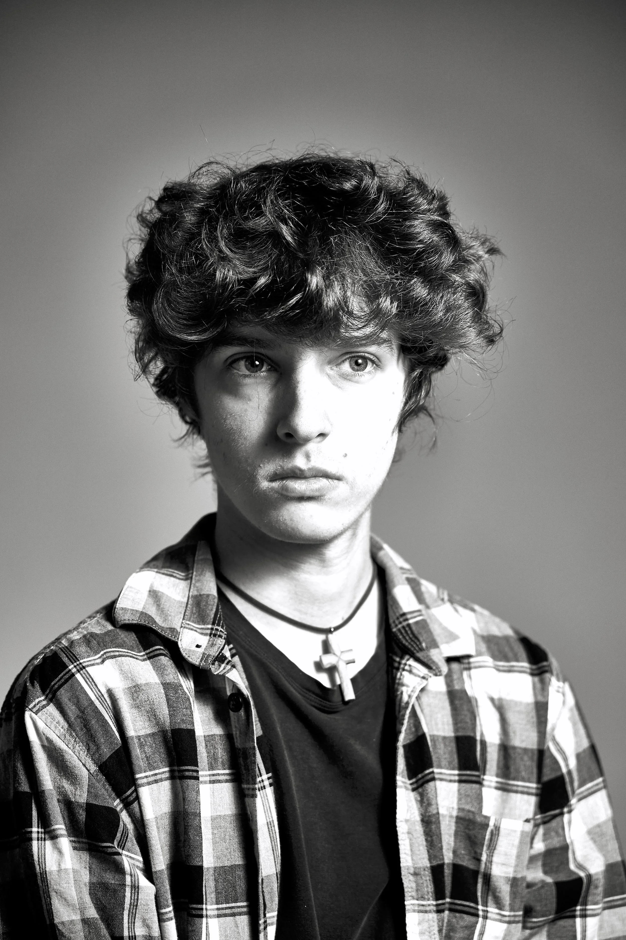 Black and white portrait of a young person with curly hair, wearing a checkered shirt and a necklace with a cross pendant.