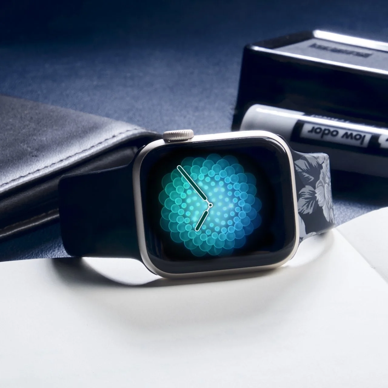 A smartwatch with a floral band displaying a digital clock on a dark background, placed on a white surface with a black wallet, a box of markers, and a notebook nearby.