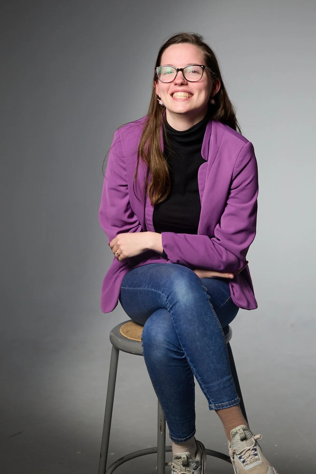 A woman with glasses and long brown hair, wearing a purple blazer over a black top, jeans, and sneakers, sitting on a stool with a neutral background.