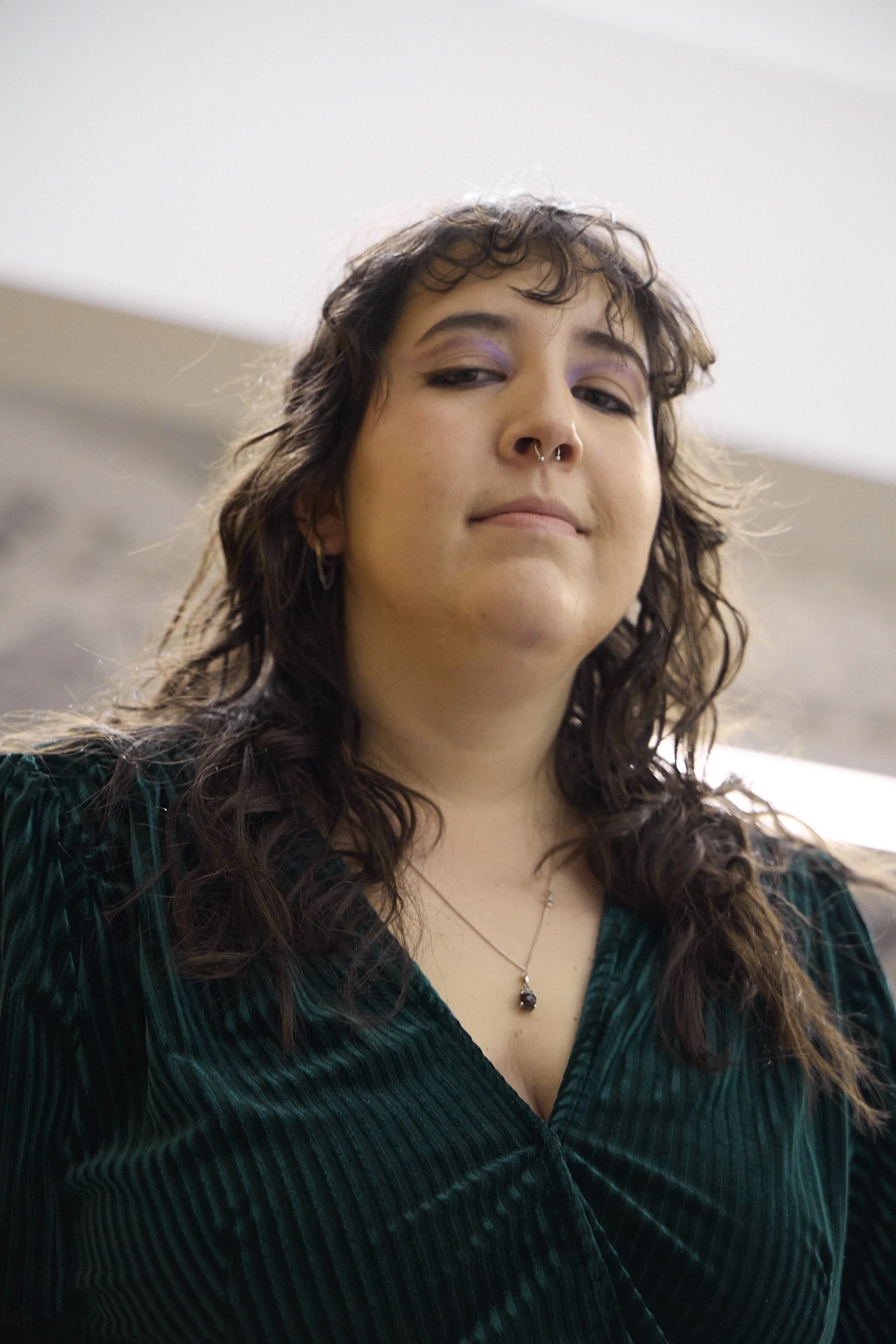 A young woman with curly brown hair, yellow eyeshadow, septum piercing, and a dark green velvet top, looking at the camera with a confident expression.