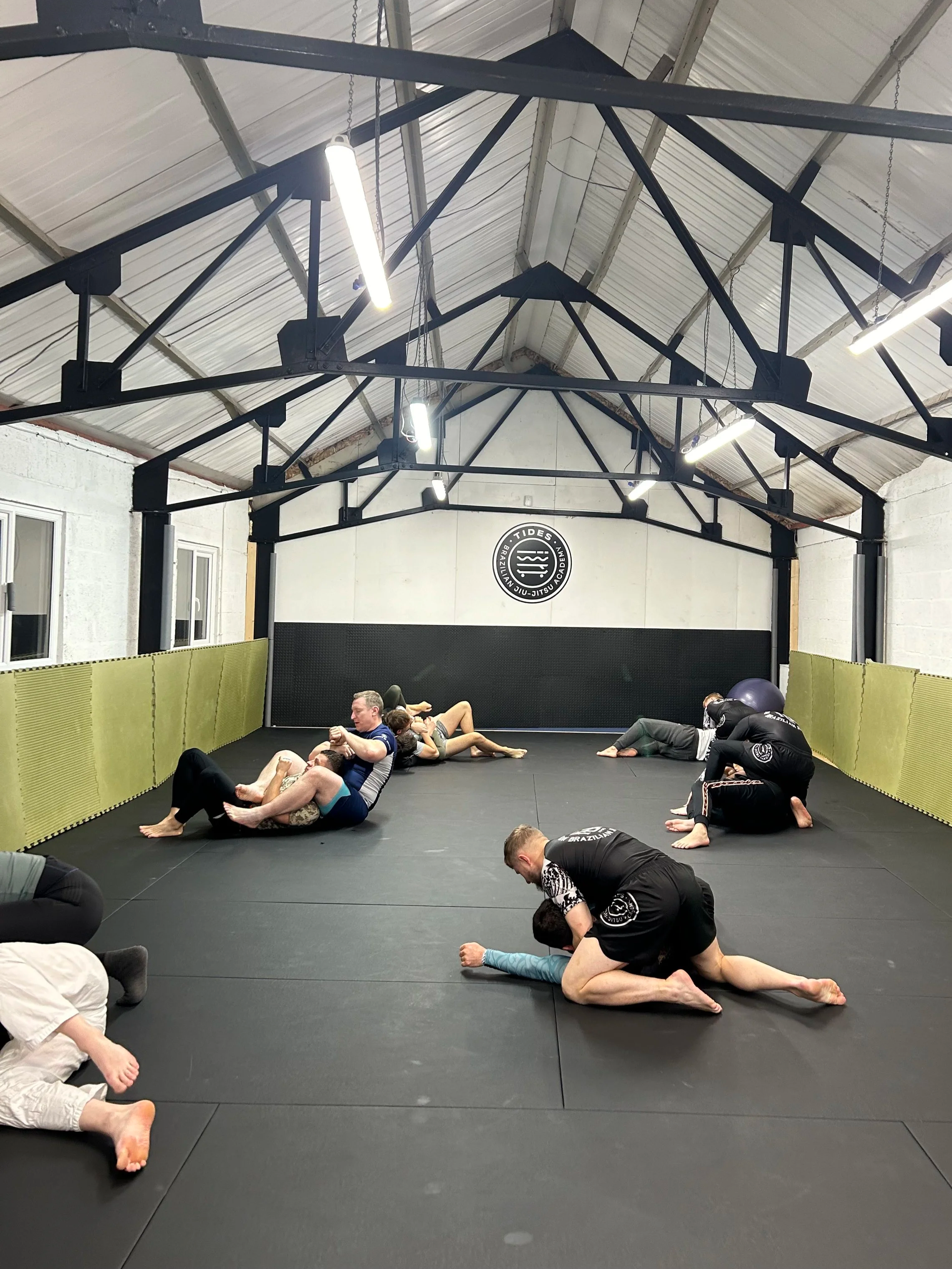 People practicing Brazilian Jiu-Jitsu on a training mat in a gym, with some grappling and others engaging in sparring.