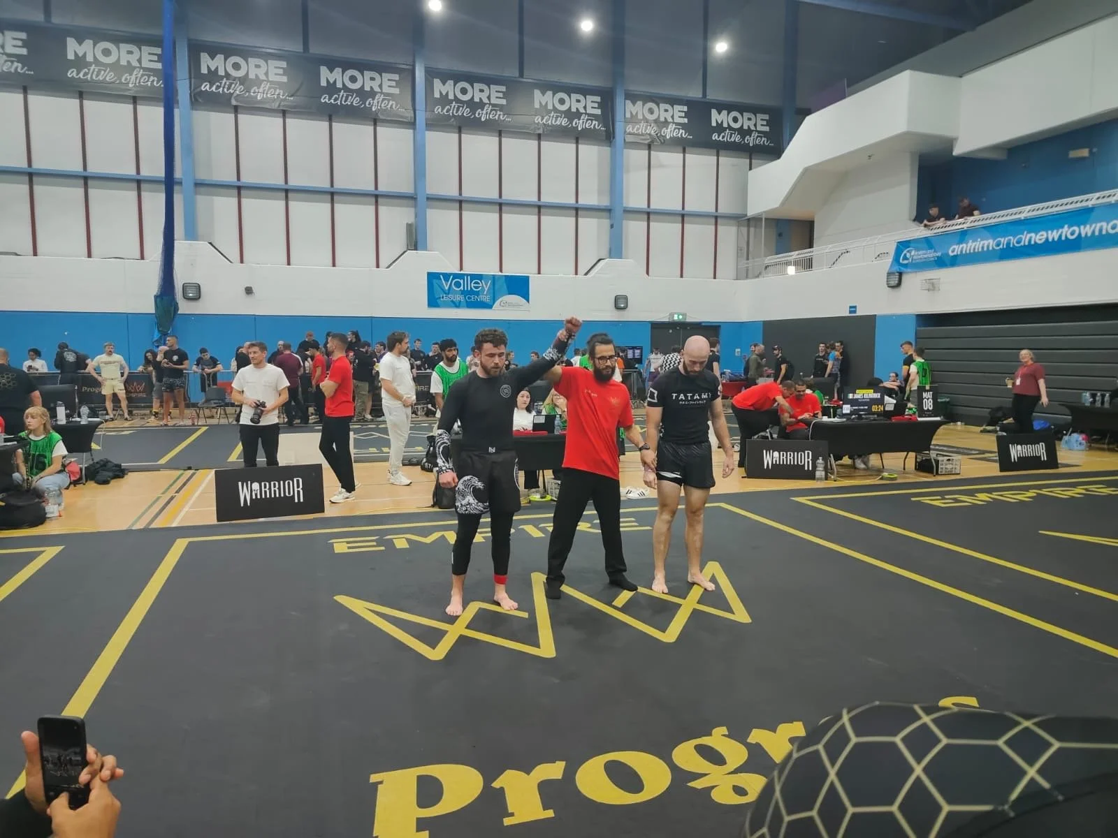 Mixed martial arts fighters and officials inside a sports facility with the word 'Progear' on the floor, spectators, and event signage in the background.