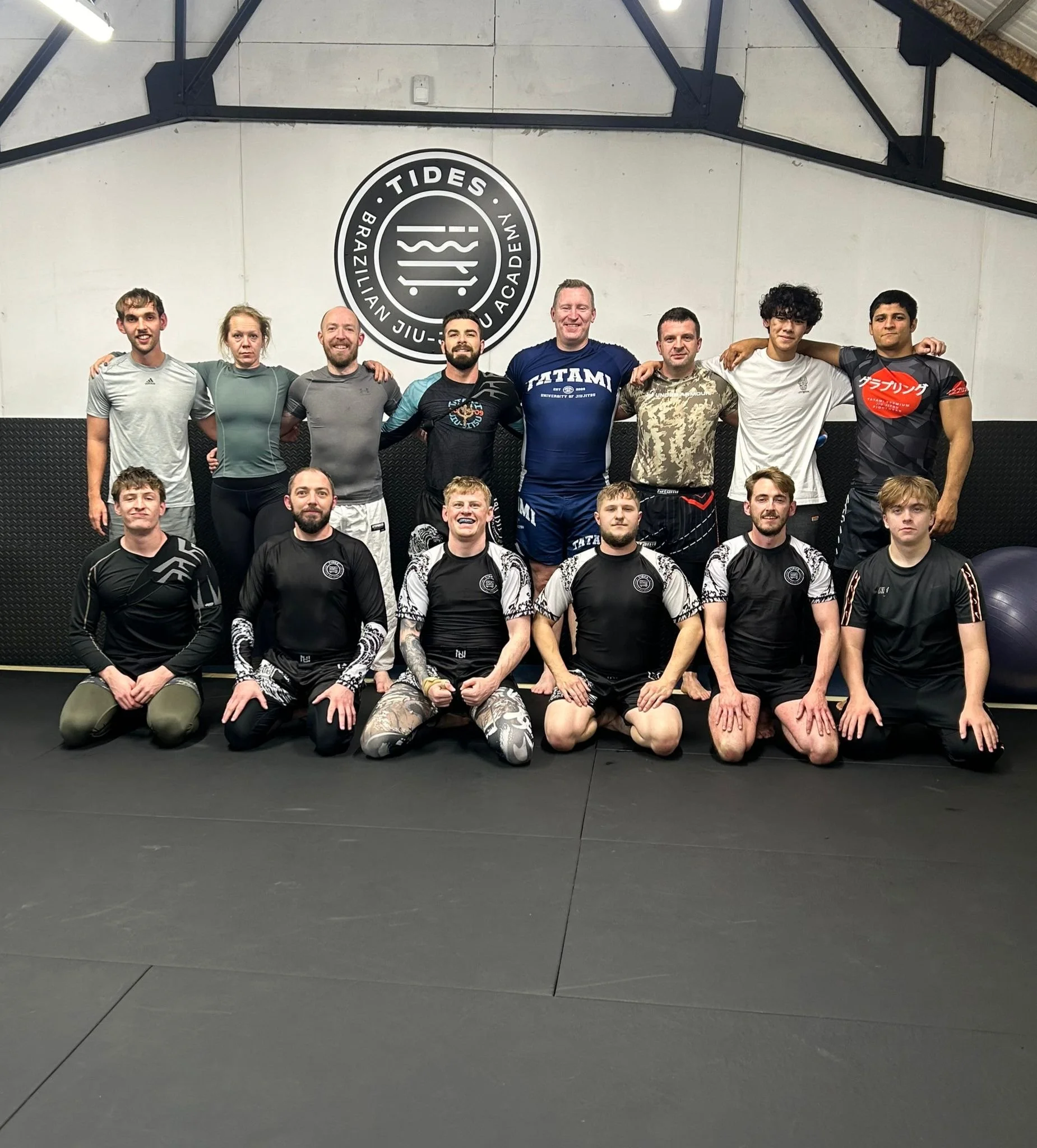 Group of people in training apparel, posing for a photo inside a martial arts gym with a sign that reads "Tides Brazilian Jiu Jitsu Academy" on the wall behind them.