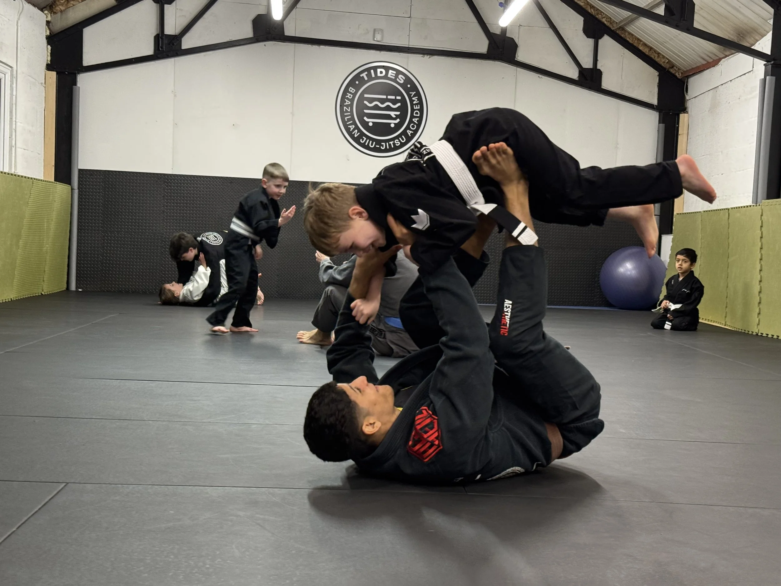 Children practicing Brazilian Jiu-Jitsu in a training gym, with at least one instructor on the mat, and a logo that reads 'Tides Brazilian Jiu-Jitsu Academy' on the wall.