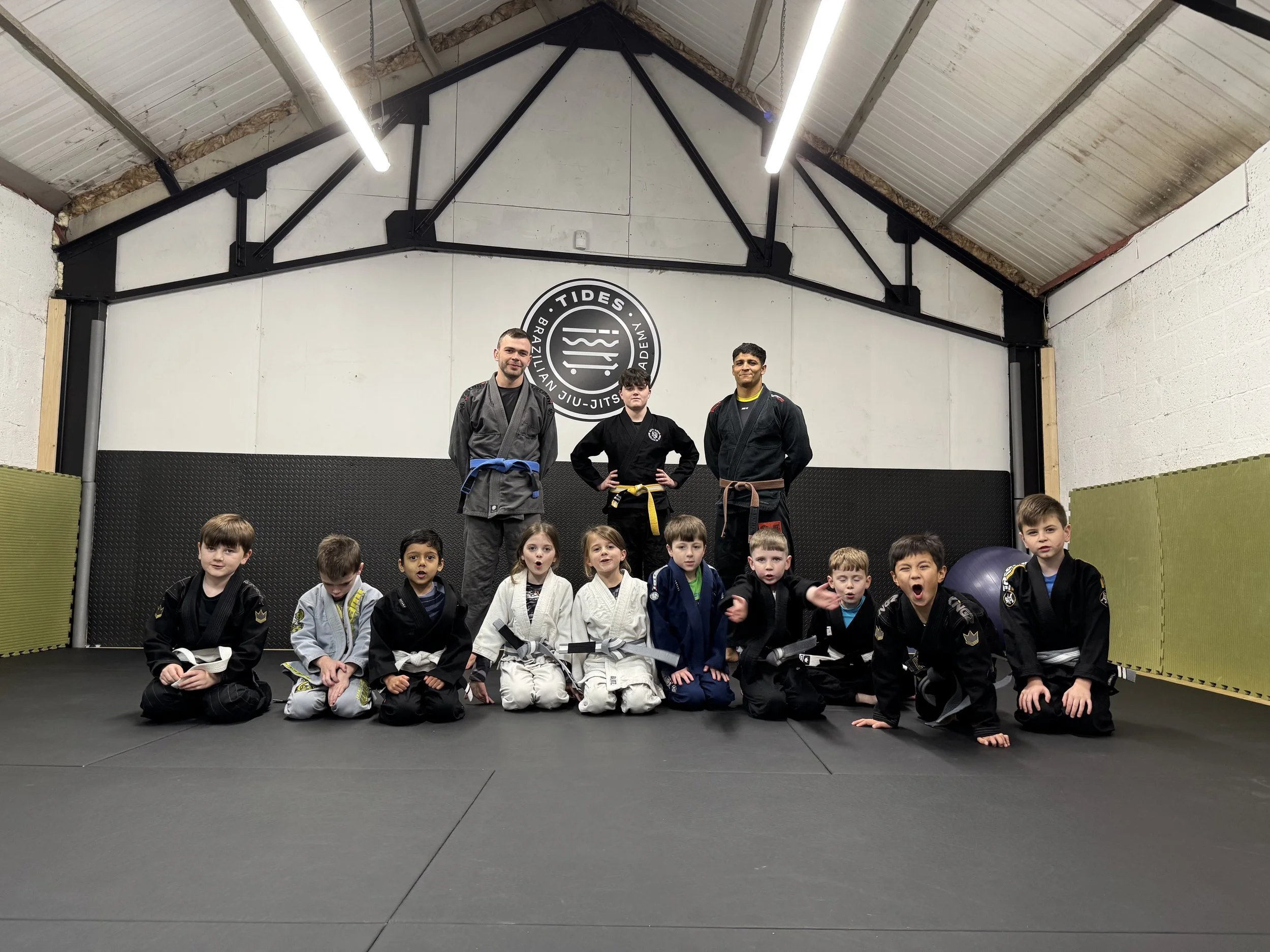 Kids and adults posing together in Brazilian Jiu-Jitsu gi uniforms at a gym, with a sign that reads "Tides Brazilian Jiu-Jitsu Academy" on the wall.
