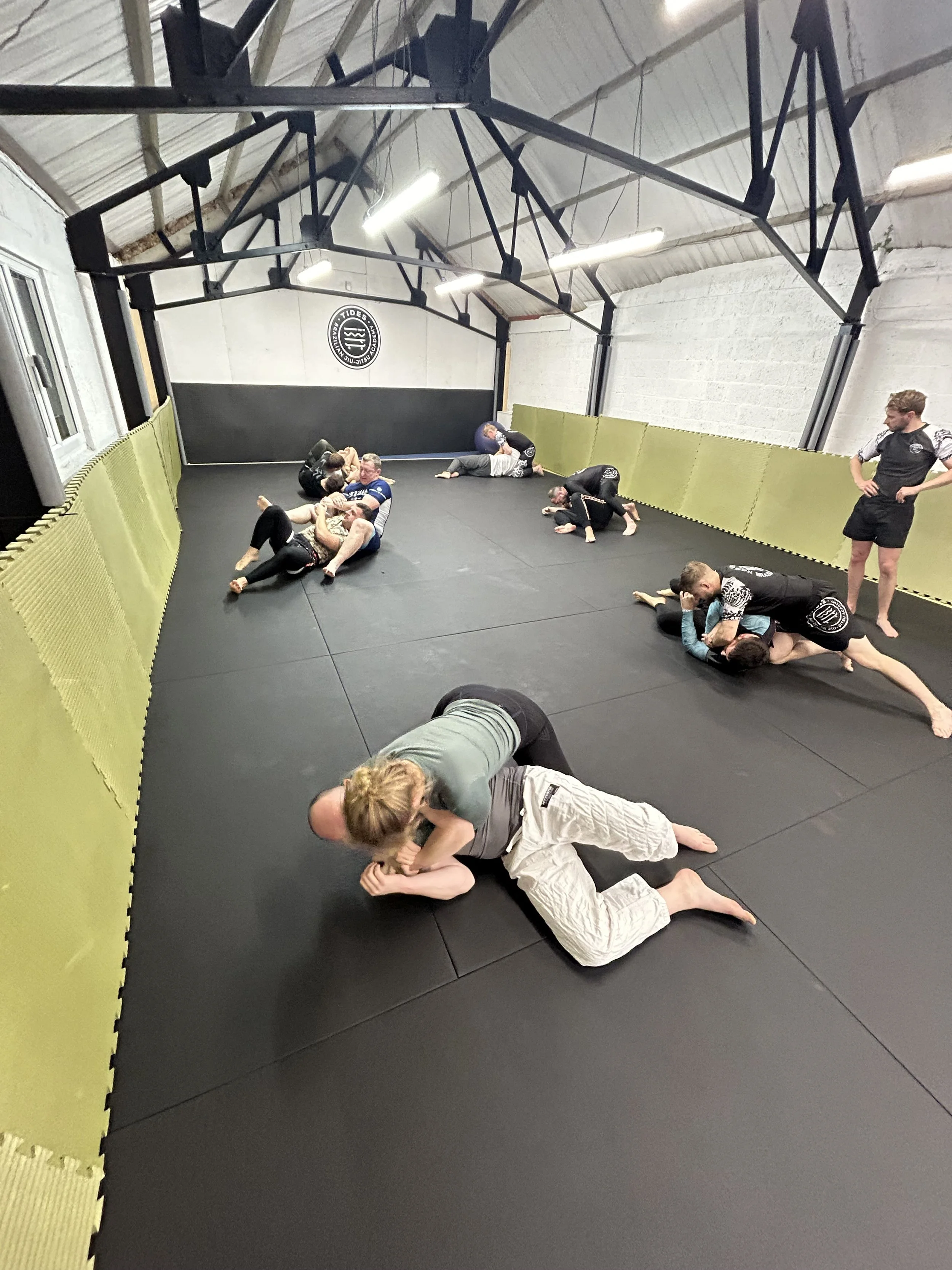 Mixed martial arts or Brazilian Jiu-Jitsu class with multiple pairs of people practicing grappling and ground fighting techniques on a padded mat in a training room, with an instructor observing.
