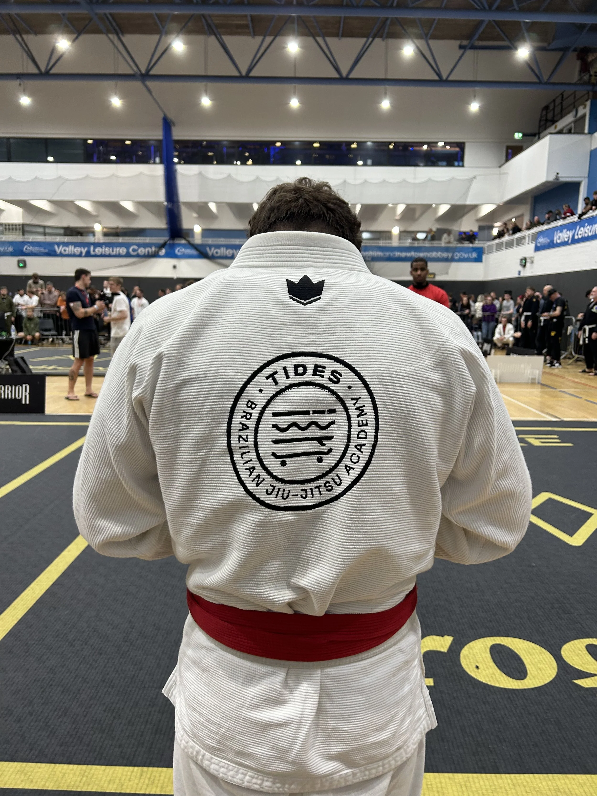 A person wearing a white martial arts gi with a red belt, standing in a sports hall. The back of the gi has a circular logo for "Tides Brazilian Jiu-Jitsu Academy" and a small black crown symbol above it. There are people and equipment in the background, indicating a martial arts event or tournament.