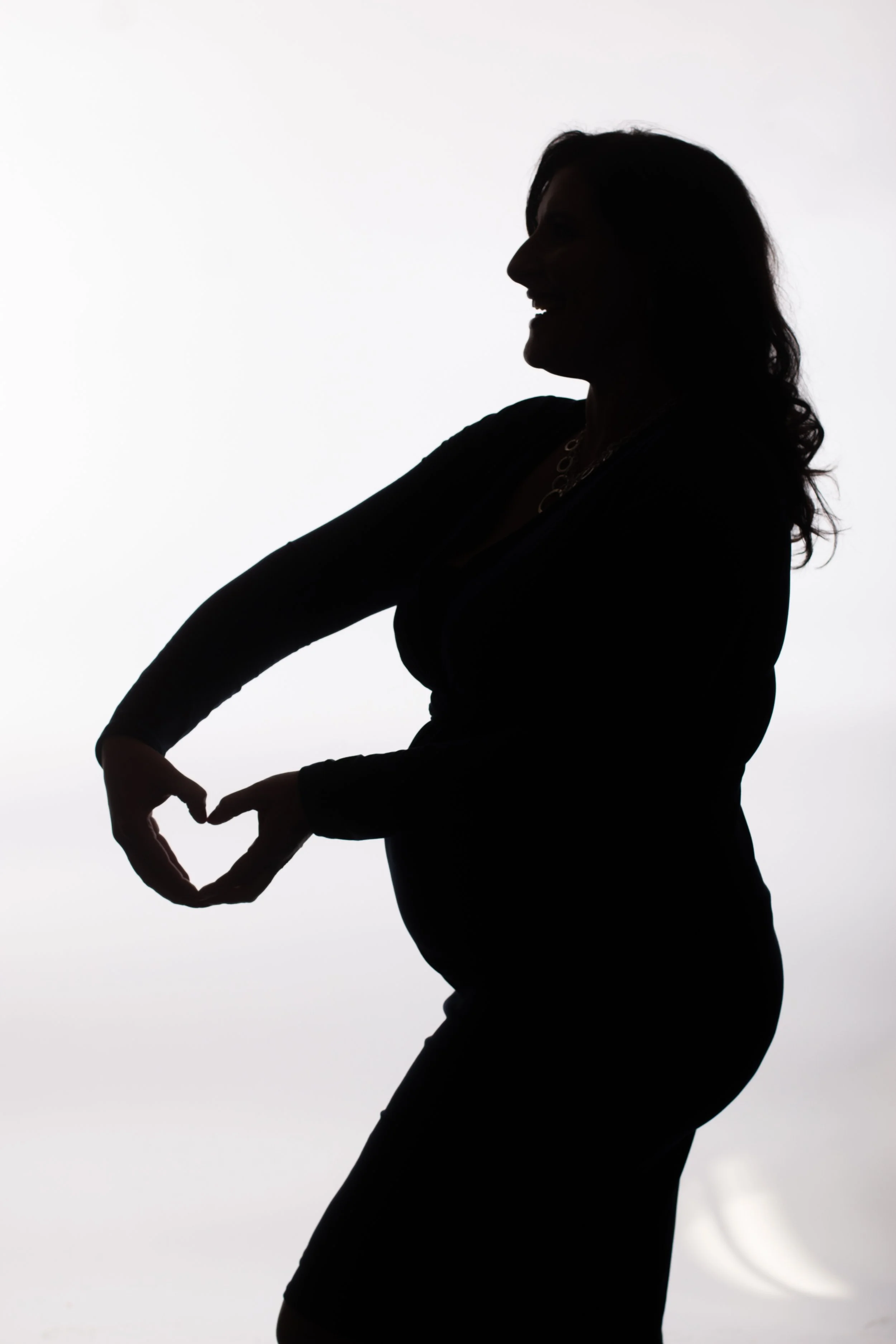Dramatic maternity silhouette of pregnant woman making a heart shape with her hands over her baby bump during an edgy studio photoshoot in Cincinnati