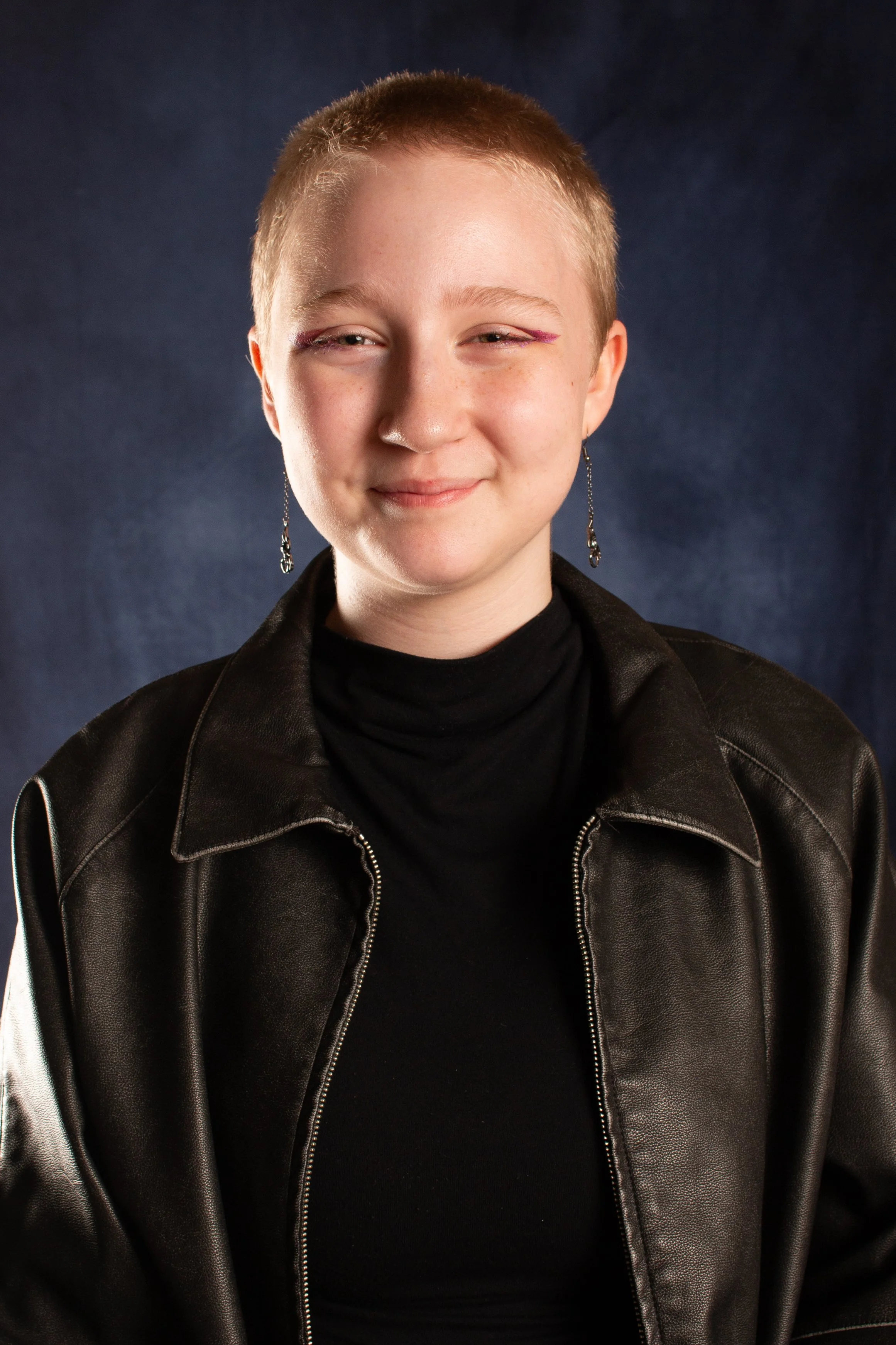  Student in black leather jacket with  shaved head smiling for school portrait  against dark backdrop 
