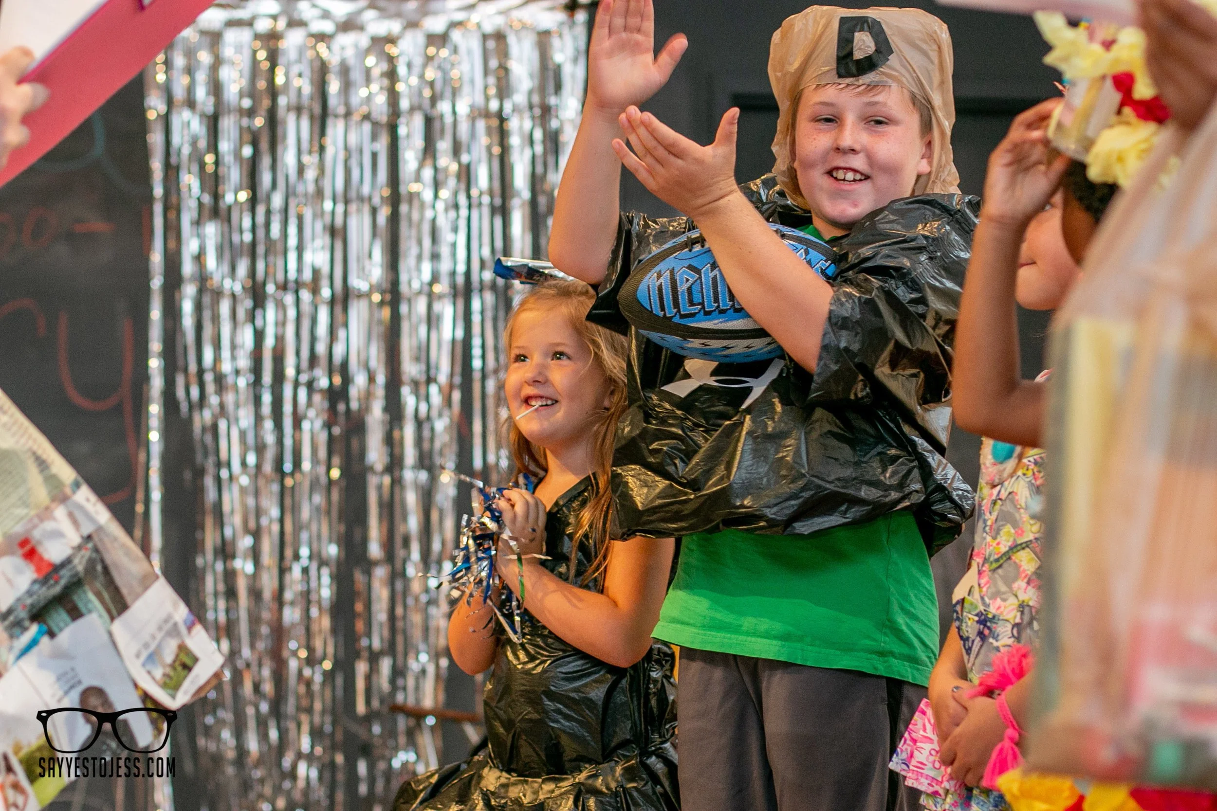 Kids waving and clapping on the trashion show runway with silver tinsel backdrop