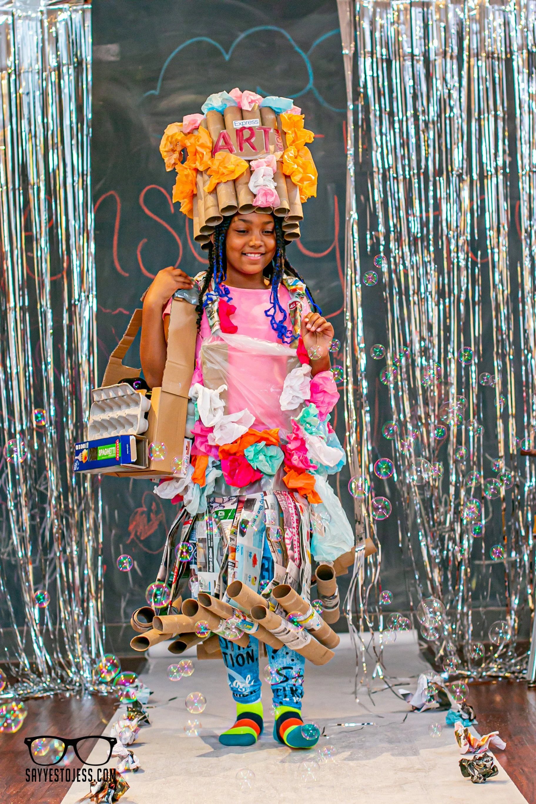 Girl in pink modeling her full recycled materials outfit with cardboard tube headpiece and bubbles on the runway at Say Yes to Jess trashion show