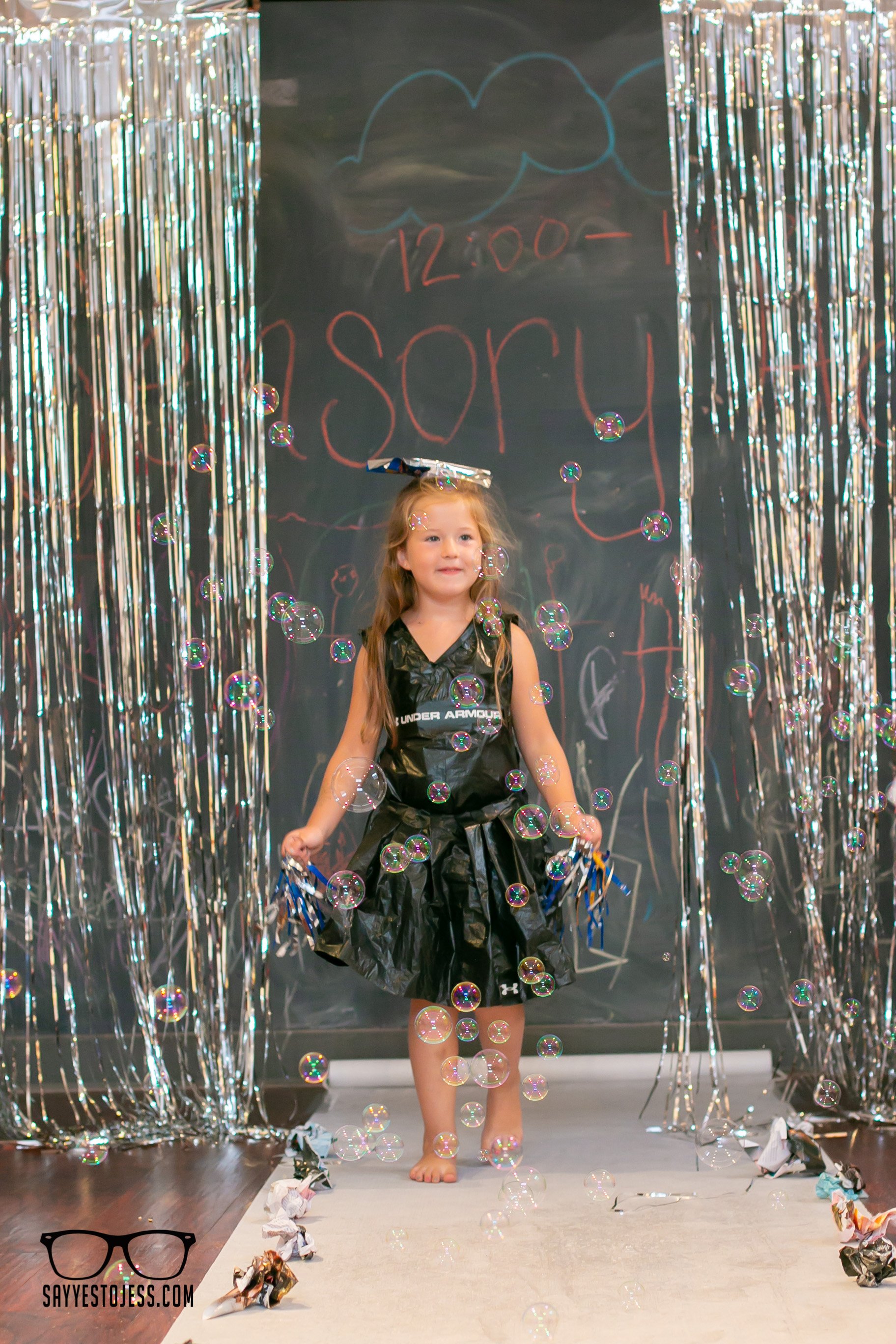 Girl in recycled trash bag dress surrounded by bubbles on the trashion show runway at Red Balloon Cafe