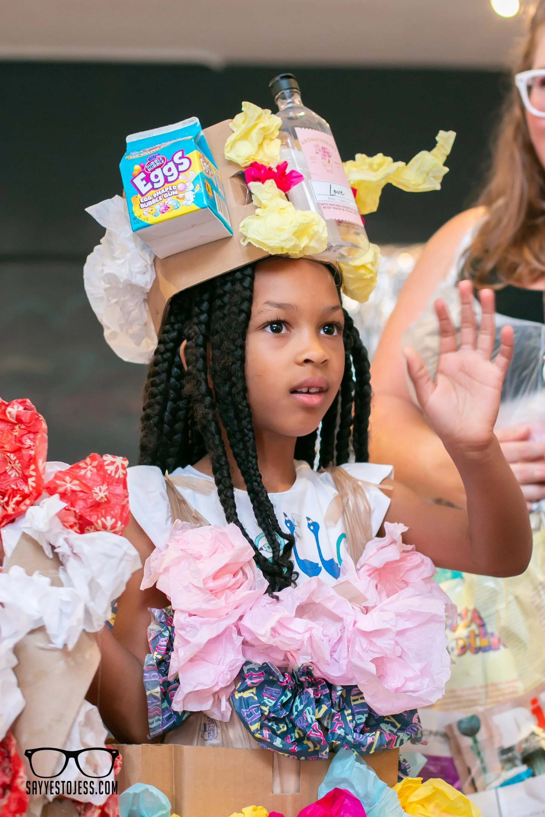 Girl with braids waving on the runway wearing a recycled paper and tissue paper outfit with creative headpiece at the Cincinnati trashion show